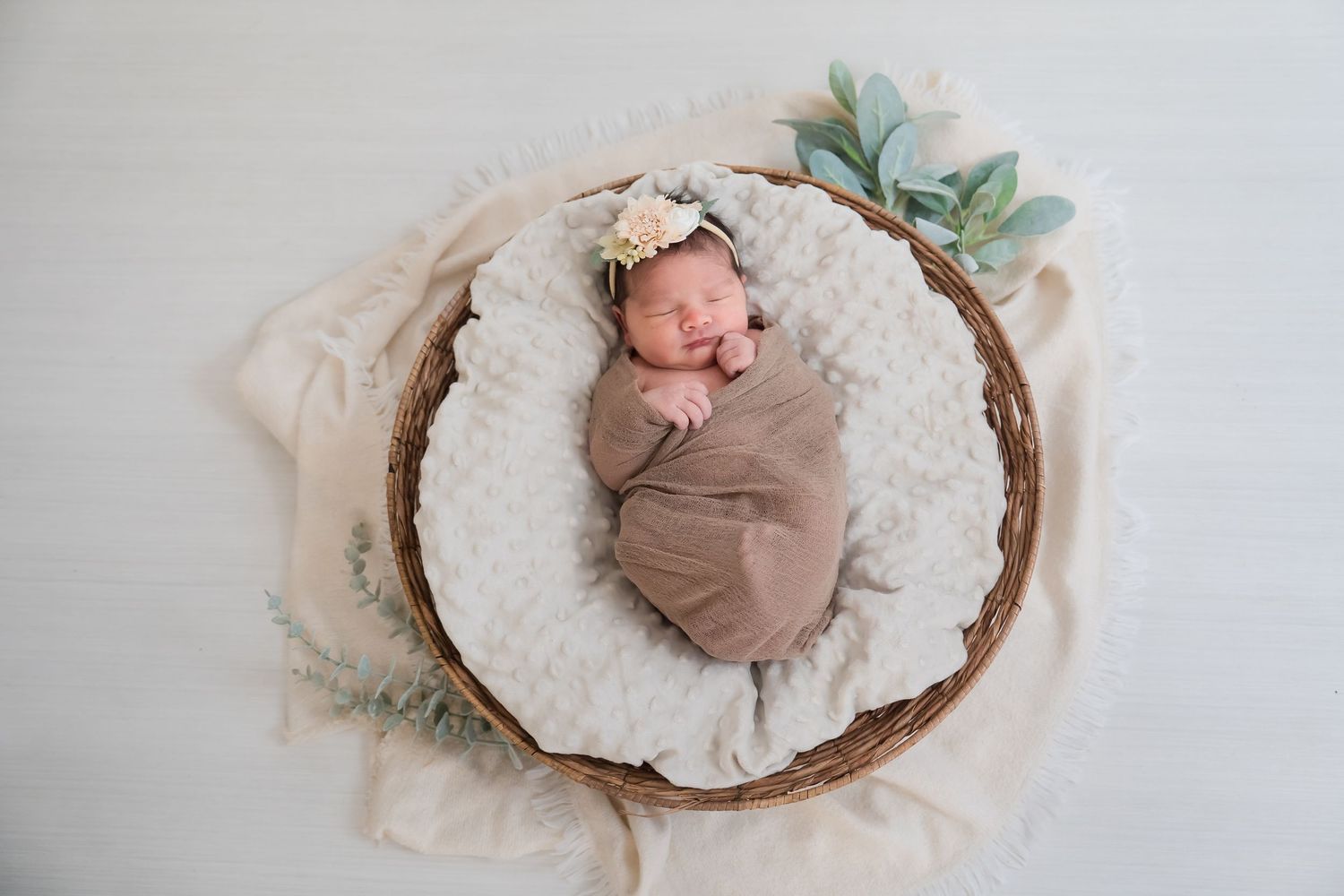 Newborn baby wrapped in brown swaddle lying on white textured blanket in wooden bowl for professional photo shoot.