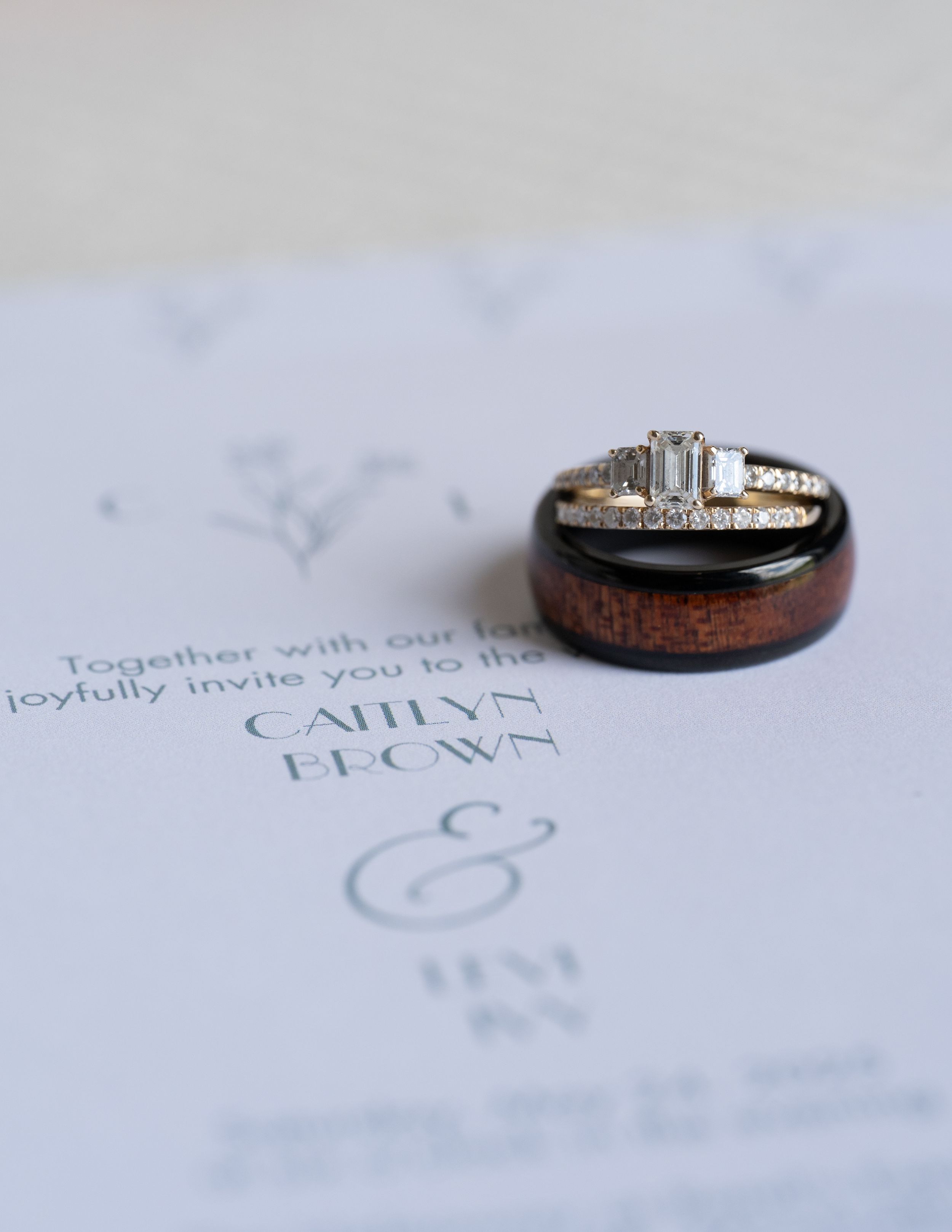 Wedding rings rest on a white invitation card with elegant text and ampersand design in soft blue ink.