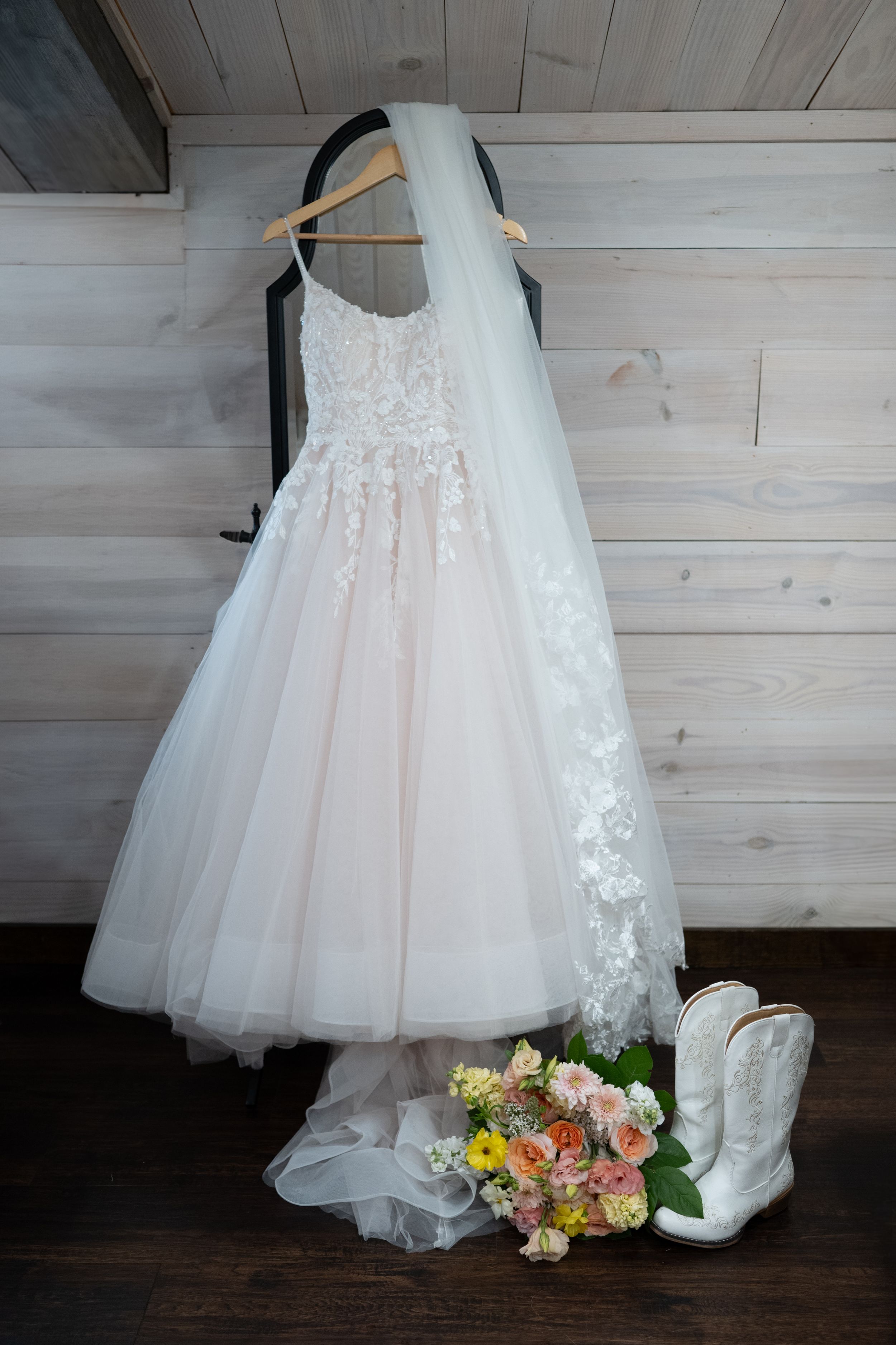 A white lace wedding dress hangs on wooden wall with bridal bouquet and cowboy boots arranged below.