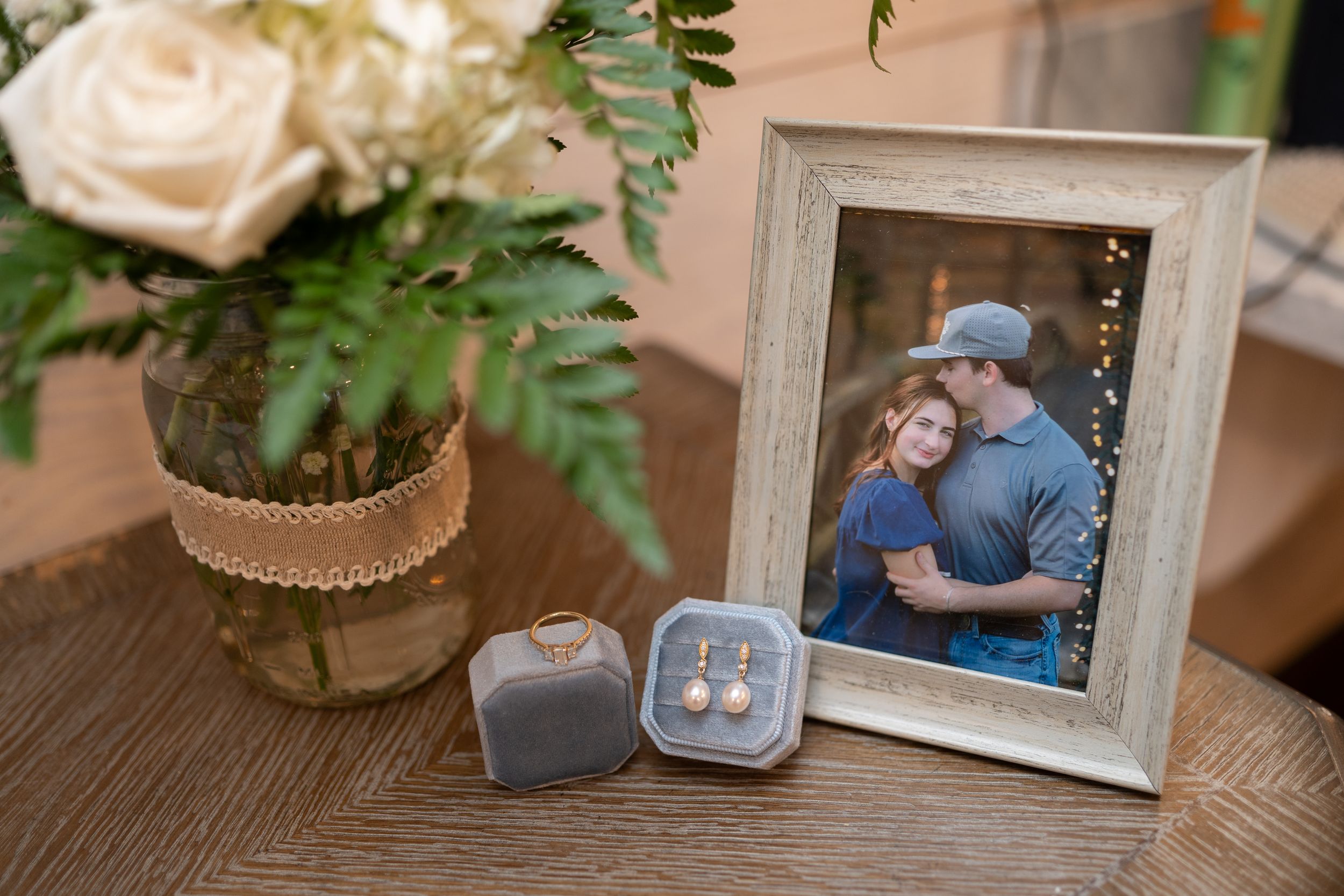 A framed photo sits on a wooden table next to a potted plant with roses and ferns in a burlap-wrapped jar and ring boxes.
