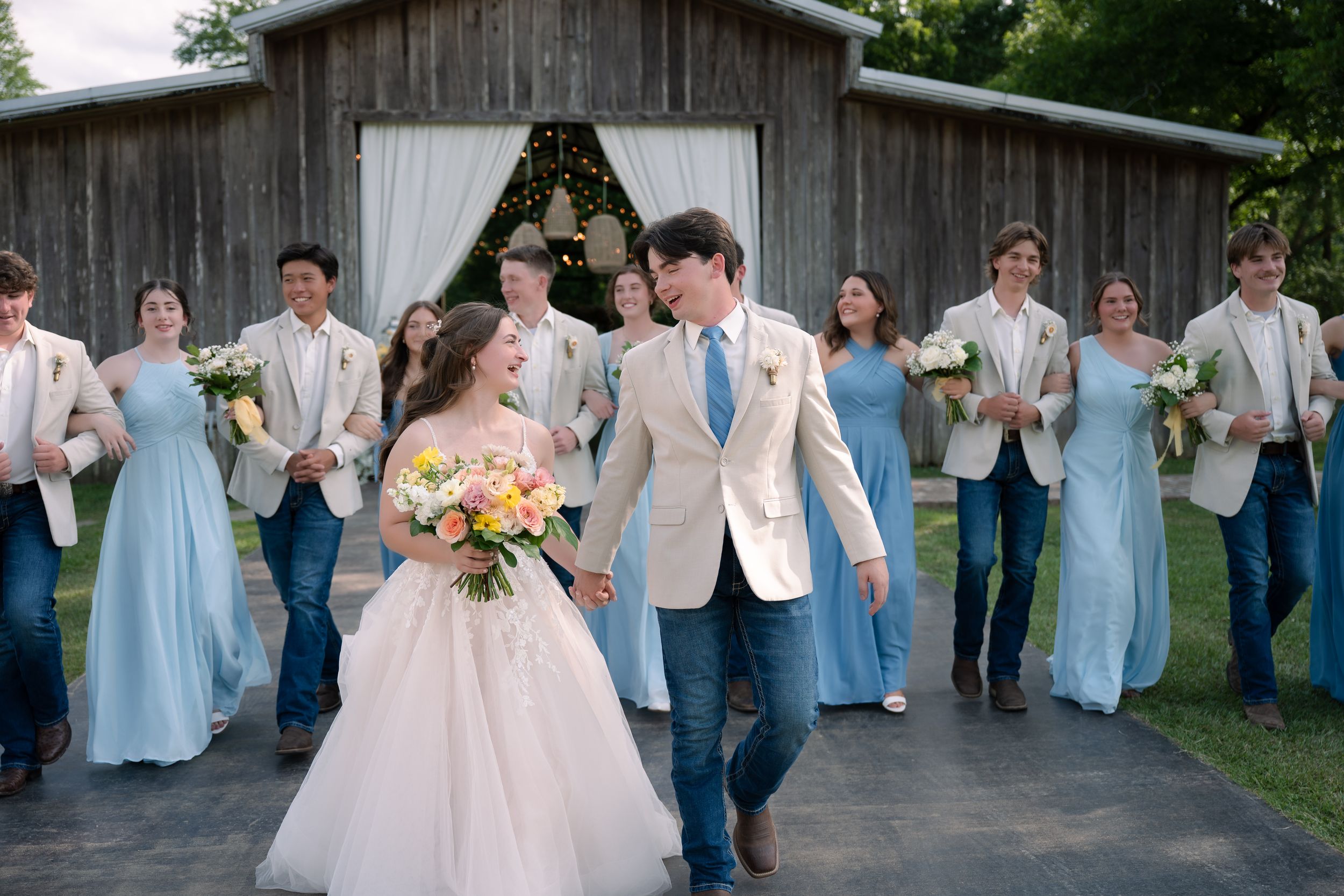 Wedding party in light blue dresses and beige suits celebrates as newlyweds share romantic moment outside rustic barn.