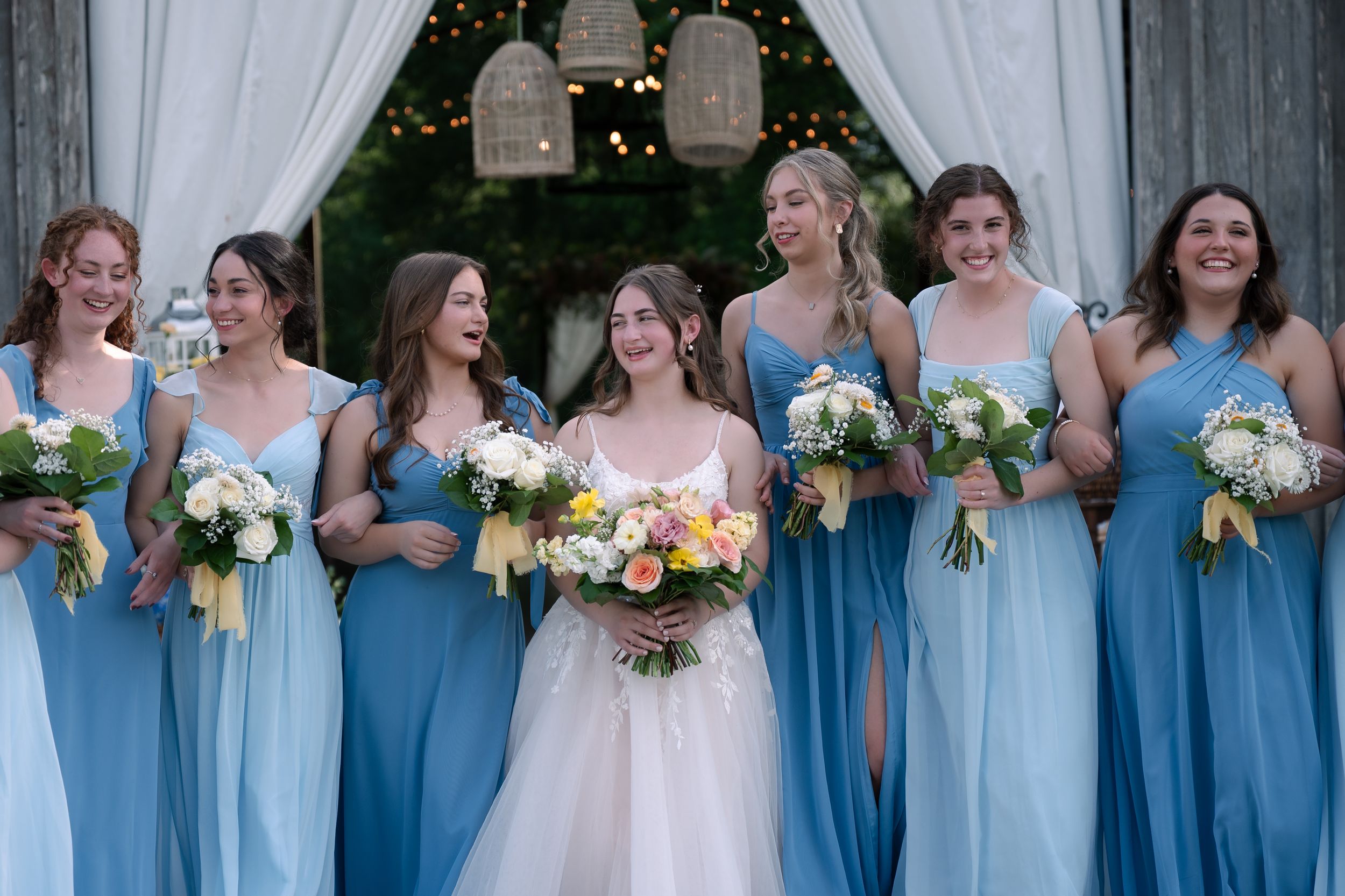 A bridal party wearing light blue bridesmaid dresses holds white and peach bouquets while standing together under an arch.