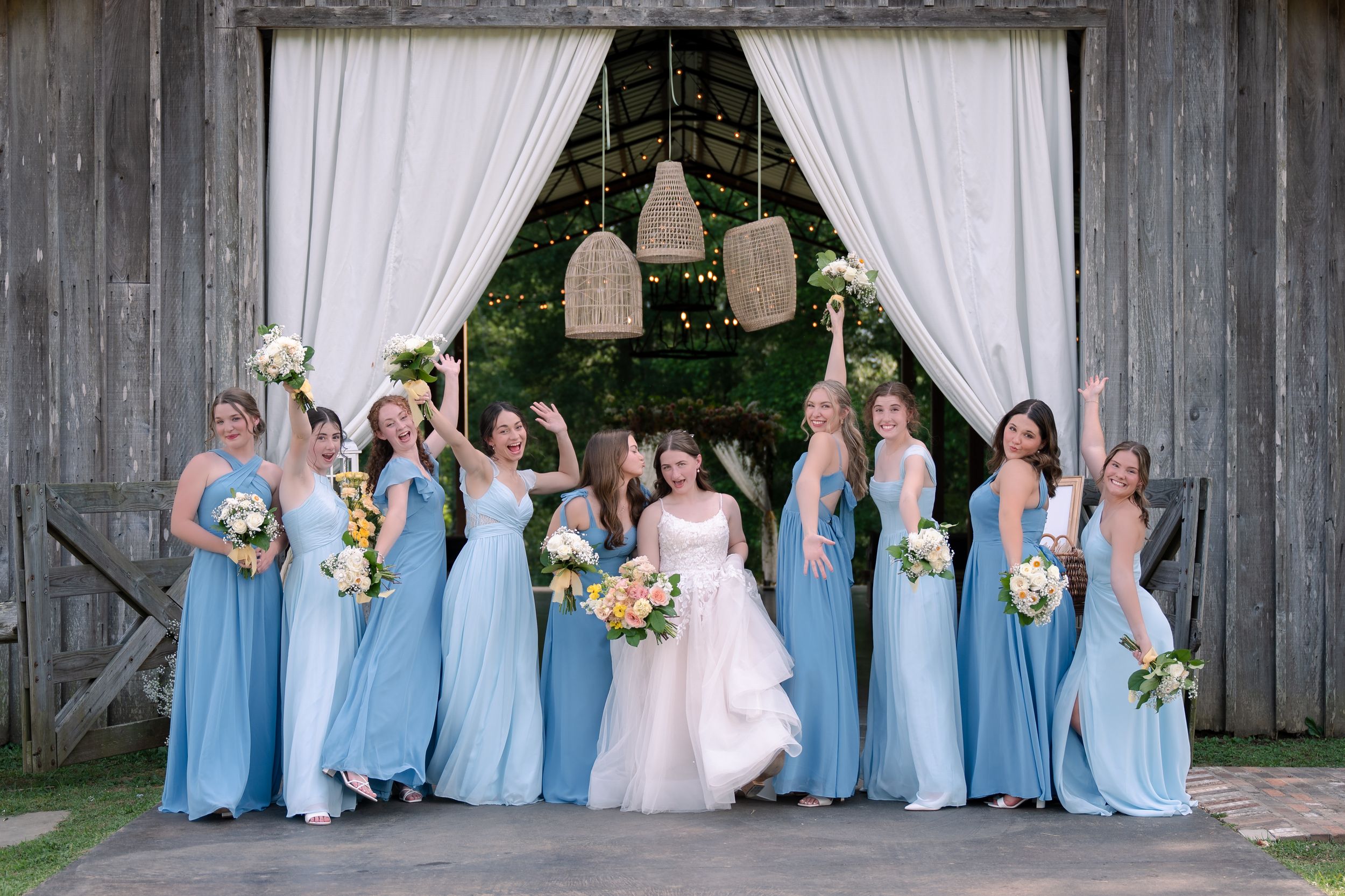 A bridal party in light blue bridesmaid dresses poses together under wooden barn entrance with white curtains and hanging lights.