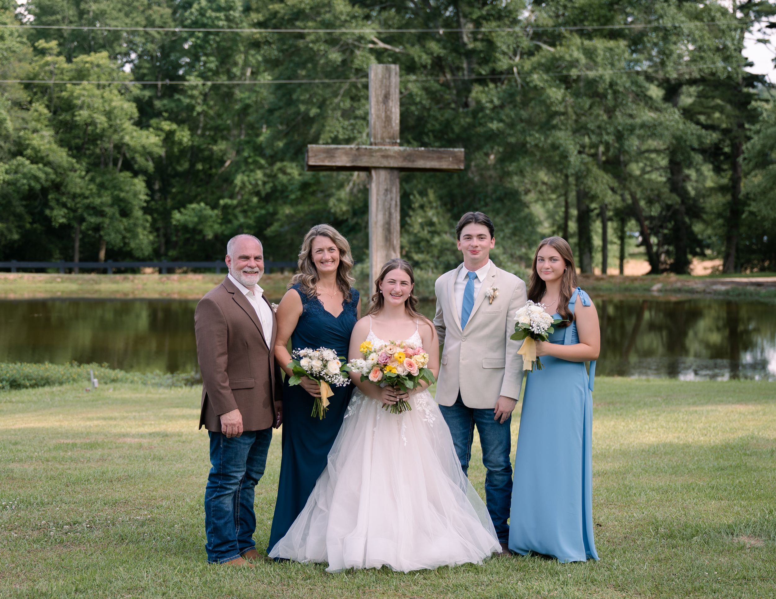 A wedding party of five people posing outdoors near a large wooden cross and pond in a grassy setting.