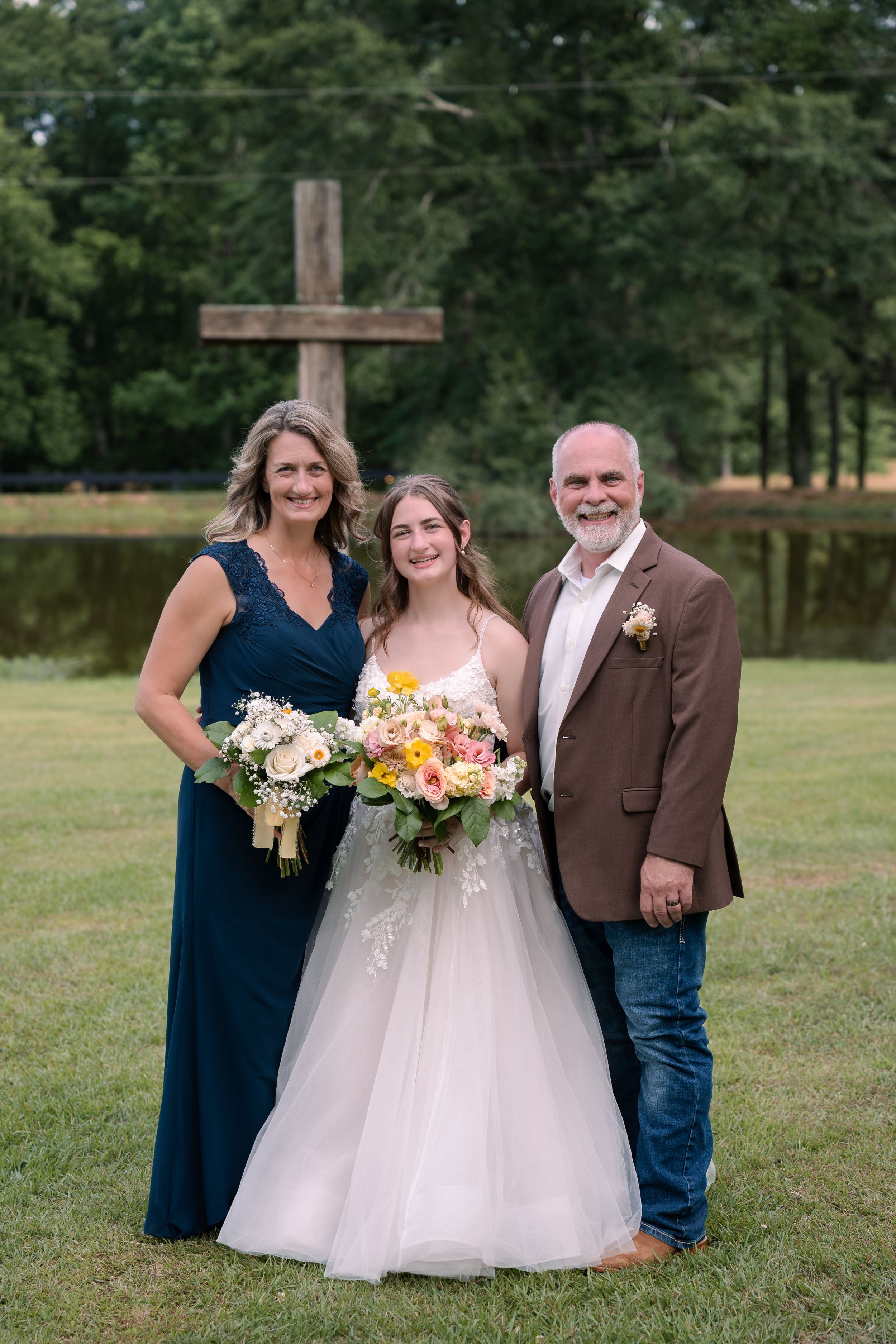Wedding photo near rustic wooden cross with bride in white gown and two others in navy and brown attire outdoors.