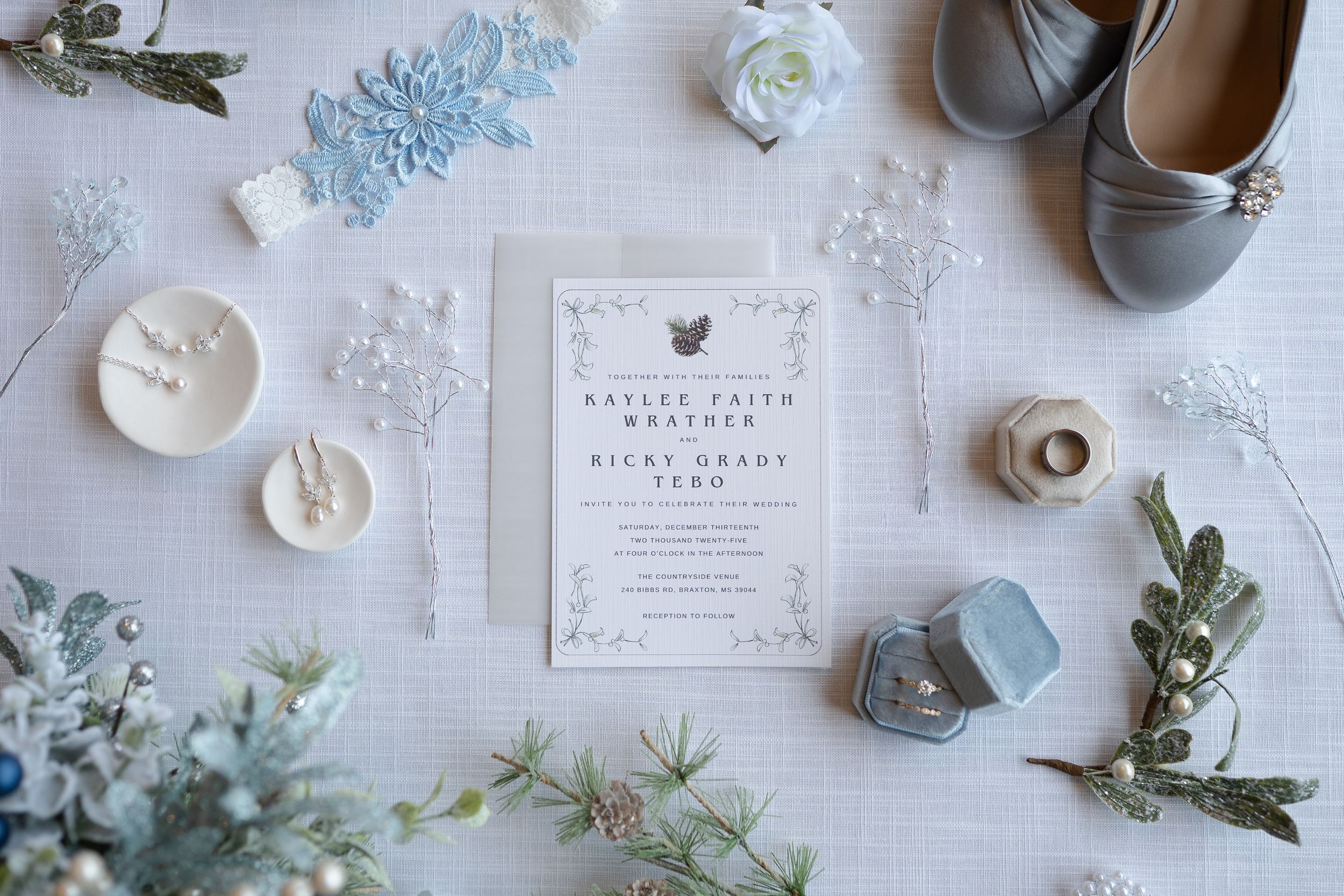 Wedding details flatlay with blue and white flowers, invitation cards, rings, and gray shoes arranged on white surface.