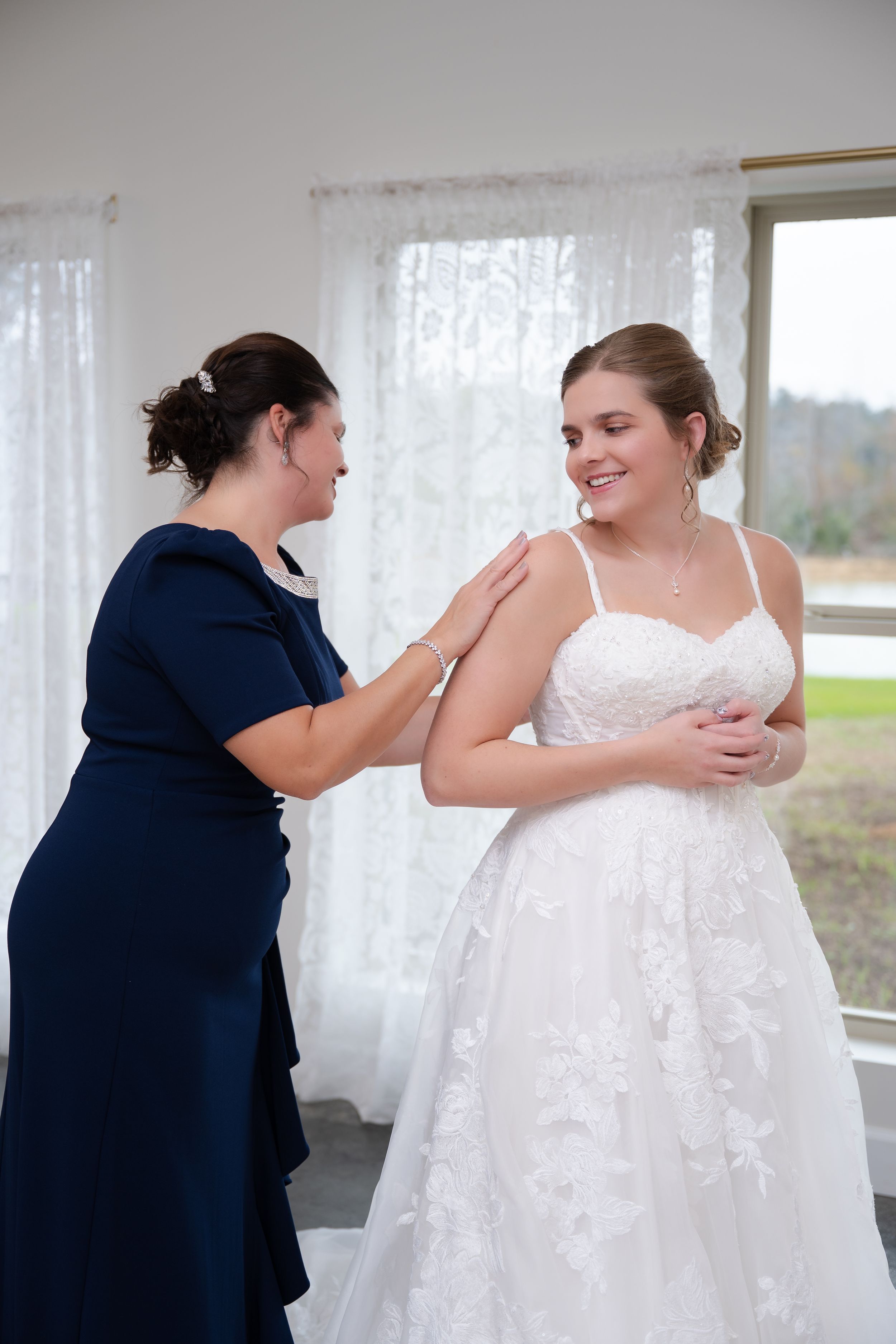 A heartwarming moment between a woman in navy blue and a bride in a white lace wedding dress by sheer curtains.