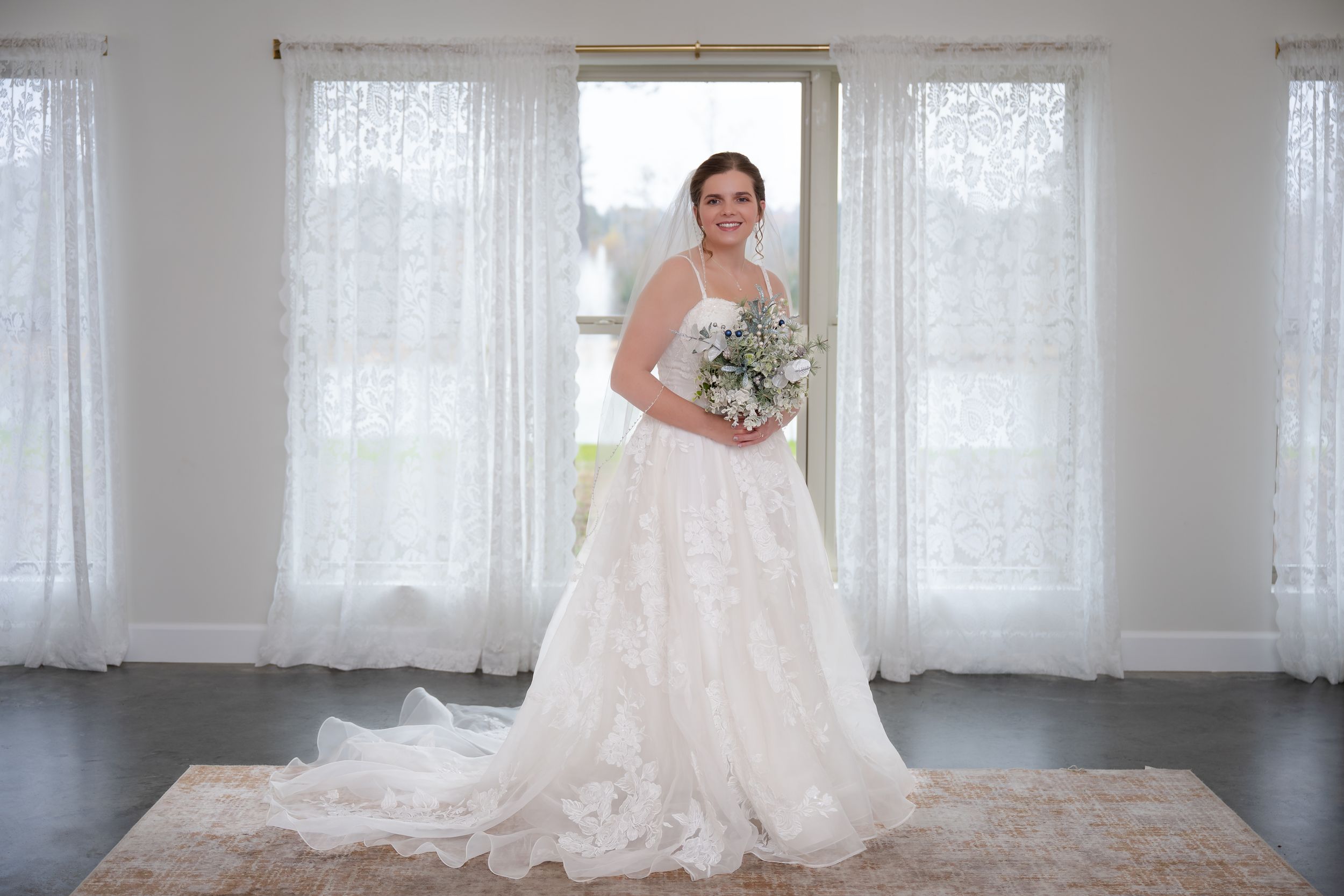 A bride in a white lace wedding gown poses in a bright sunlit room with sheer curtains.
