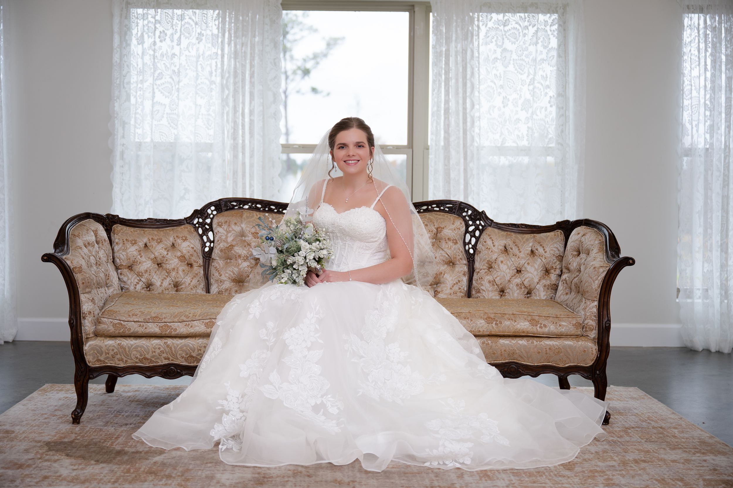 A bride in a white ballgown wedding dress poses elegantly on an antique beige sofa in a bright sunlit room.