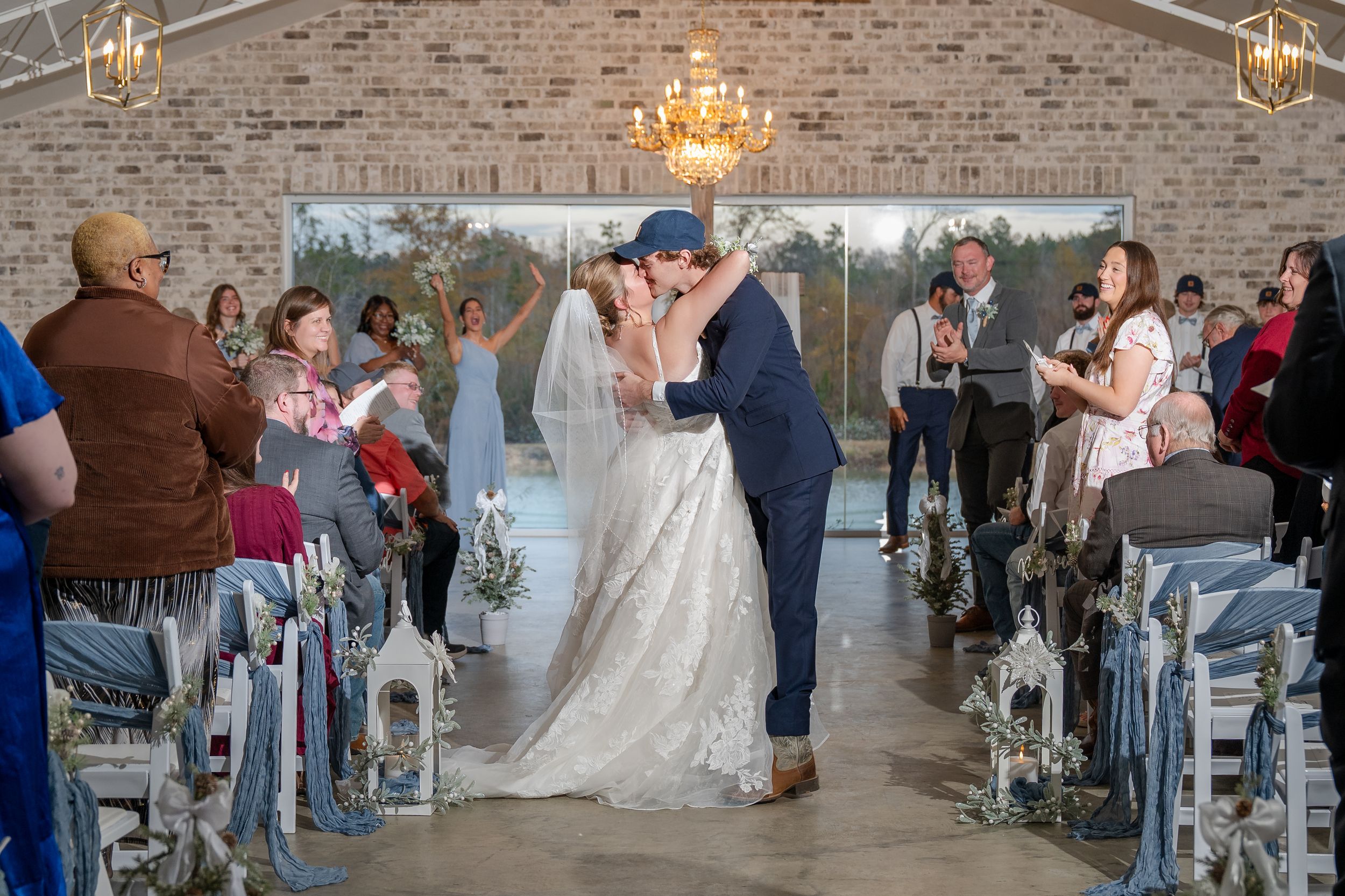 Newlyweds share their first kiss at a rustic indoor ceremony with brick walls and chandeliers.