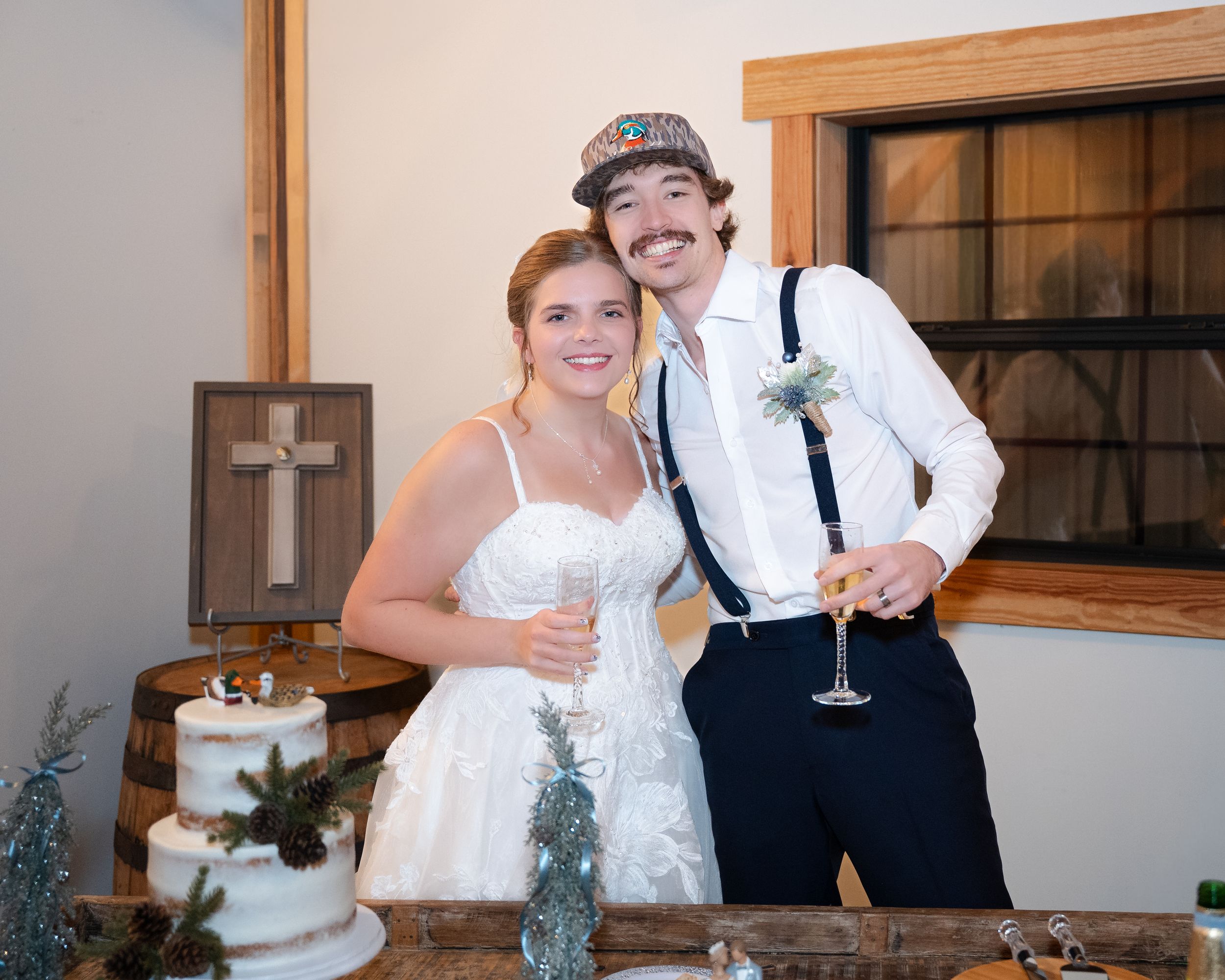 Newlywed couple cuts their rustic white wedding cake at a barn venue reception while holding champagne glasses.