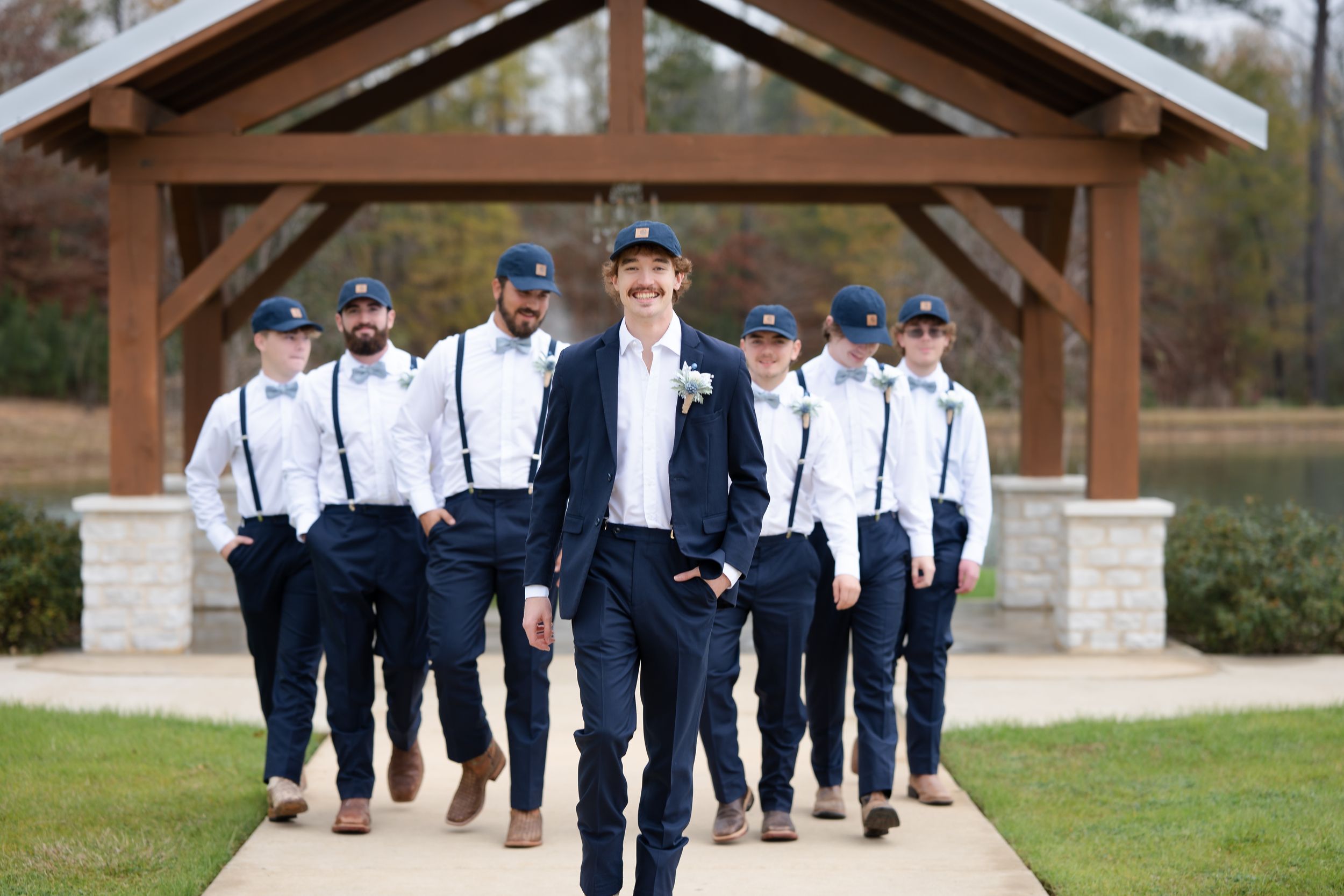 Wedding party in navy blue suits walks through a rustic wooden gazebo during an outdoor celebration.