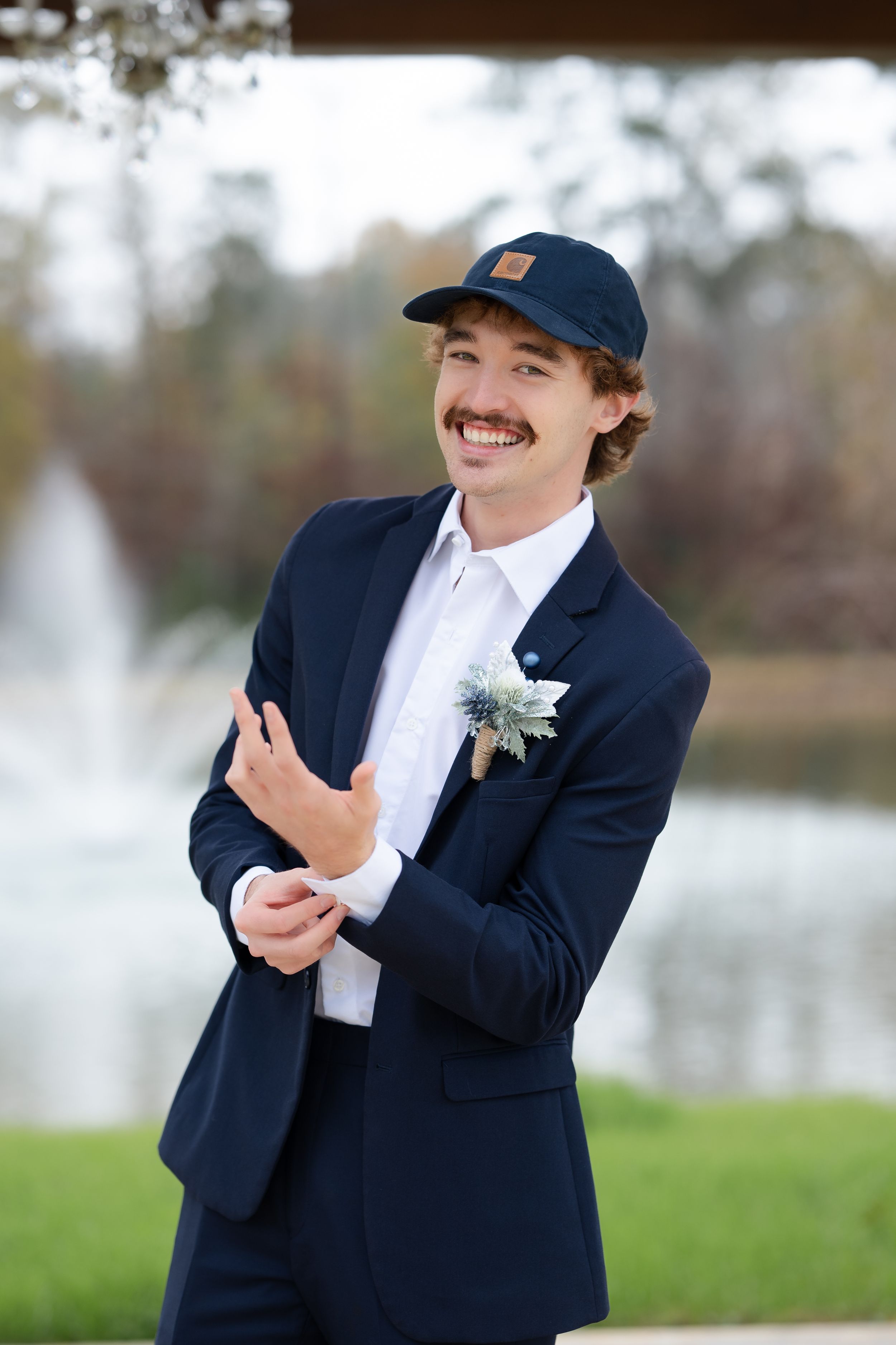 A person in a navy suit and baseball cap laughs joyfully while wearing a boutonniere at an outdoor event.
