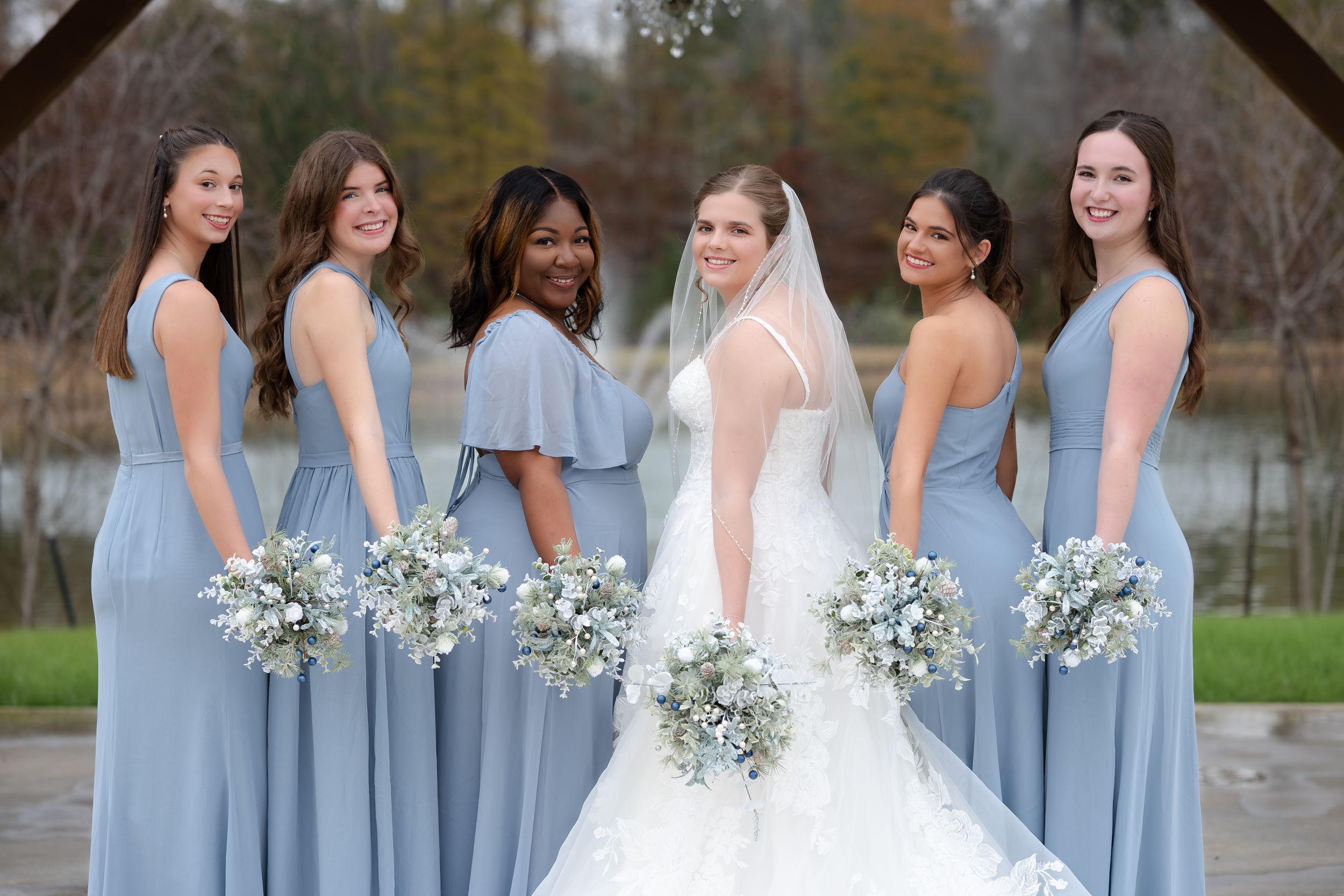 Wedding party in light blue bridesmaid dresses posing with bride in white gown by a lake.