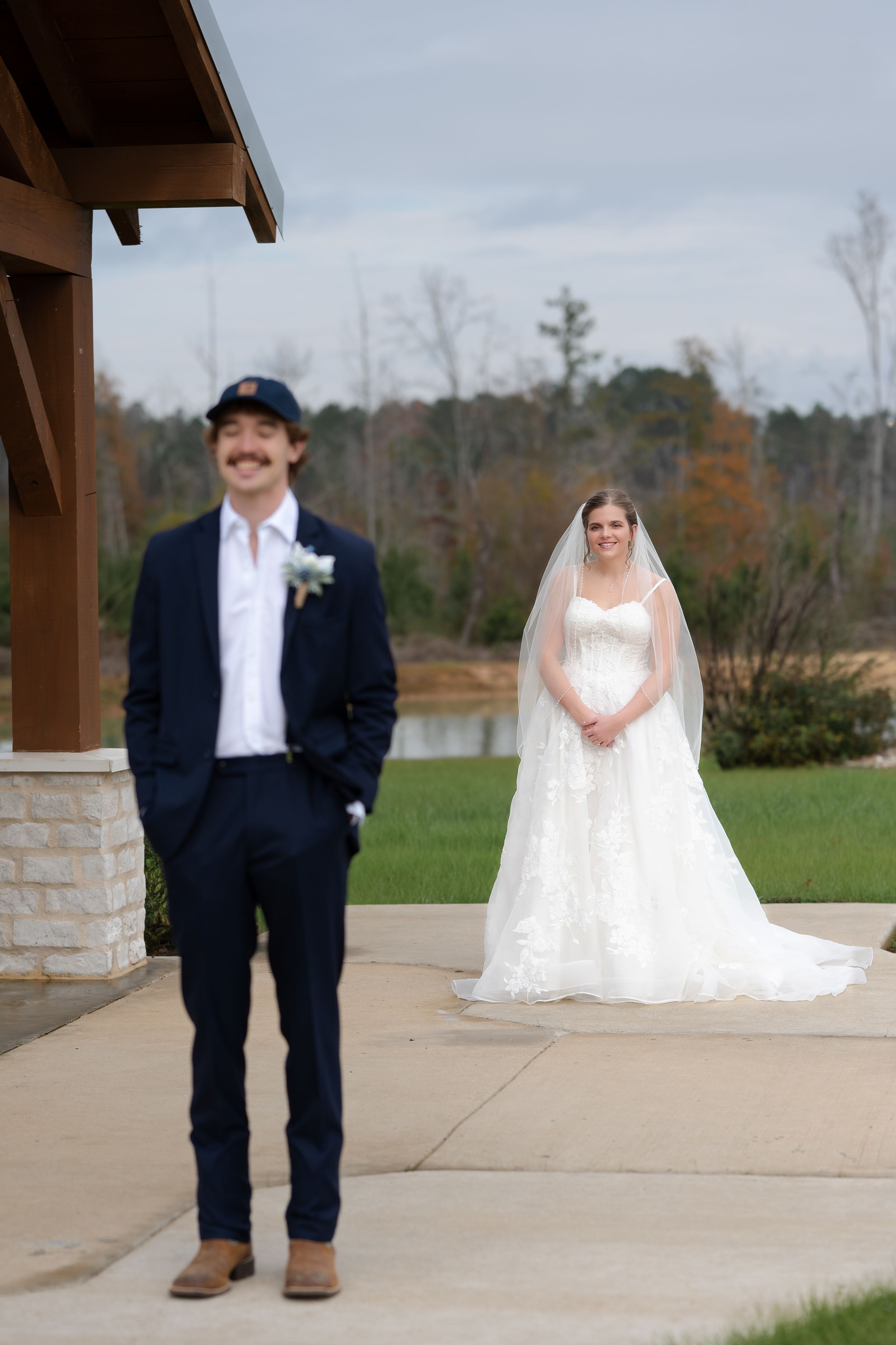 First look wedding photo with couple standing apart on concrete patio with autumn trees in background.