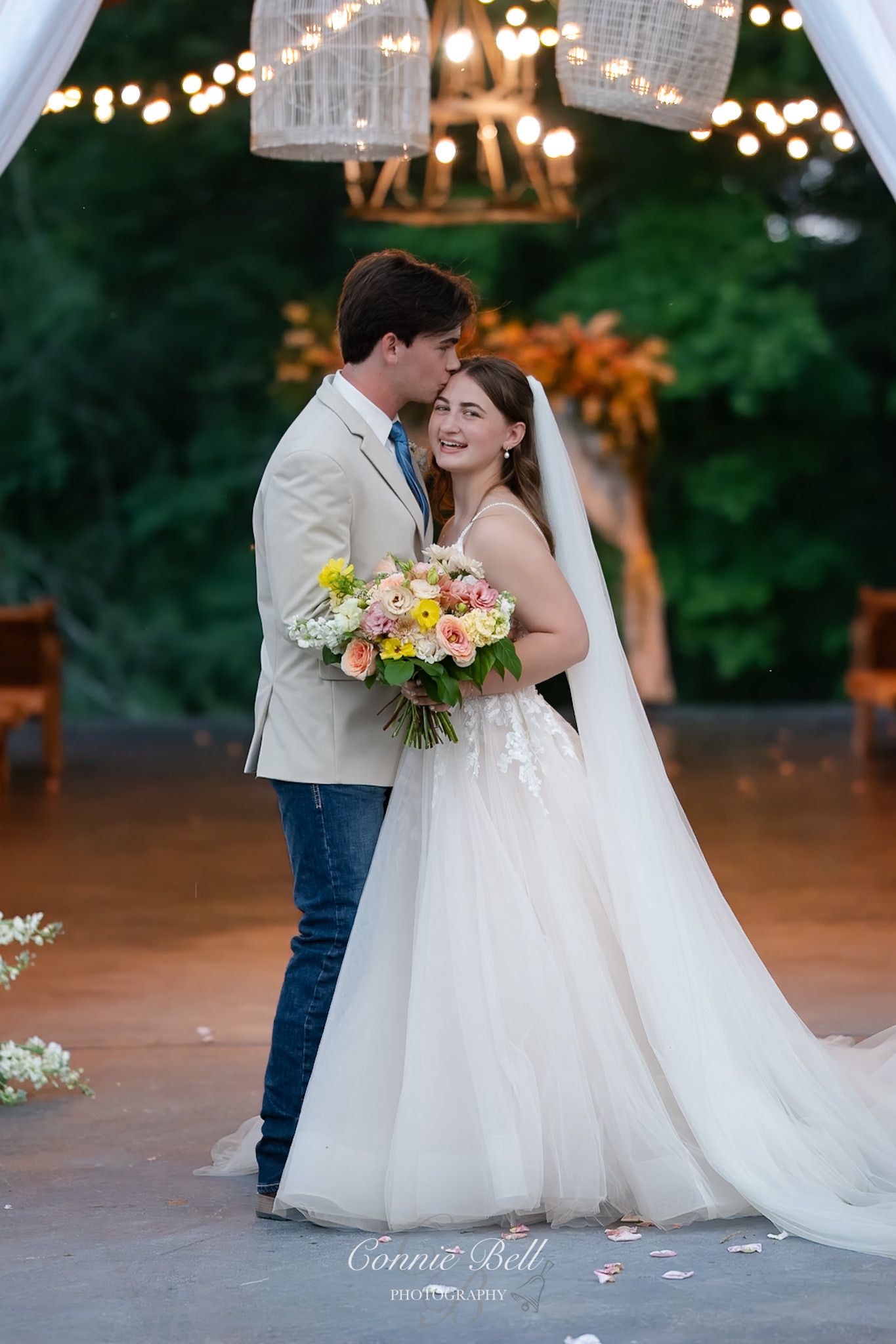 A couple celebrates their wedding day under twinkling lights, with the bride holding a vibrant yellow and coral floral bouquet.