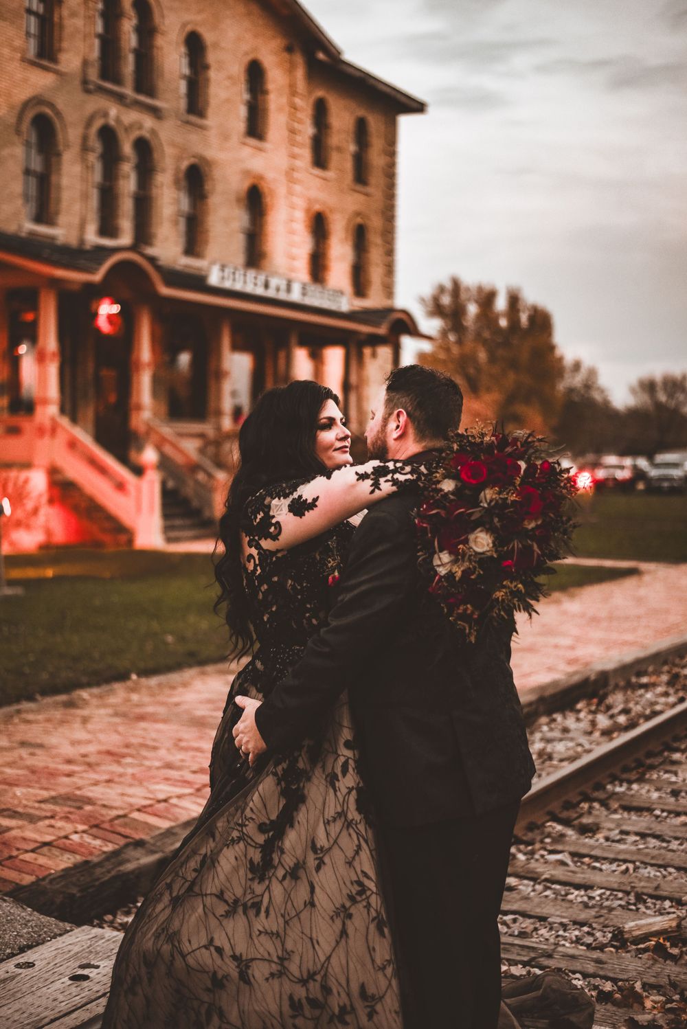 Elegant couple in formal attire embrace near vintage brick building and railway tracks.