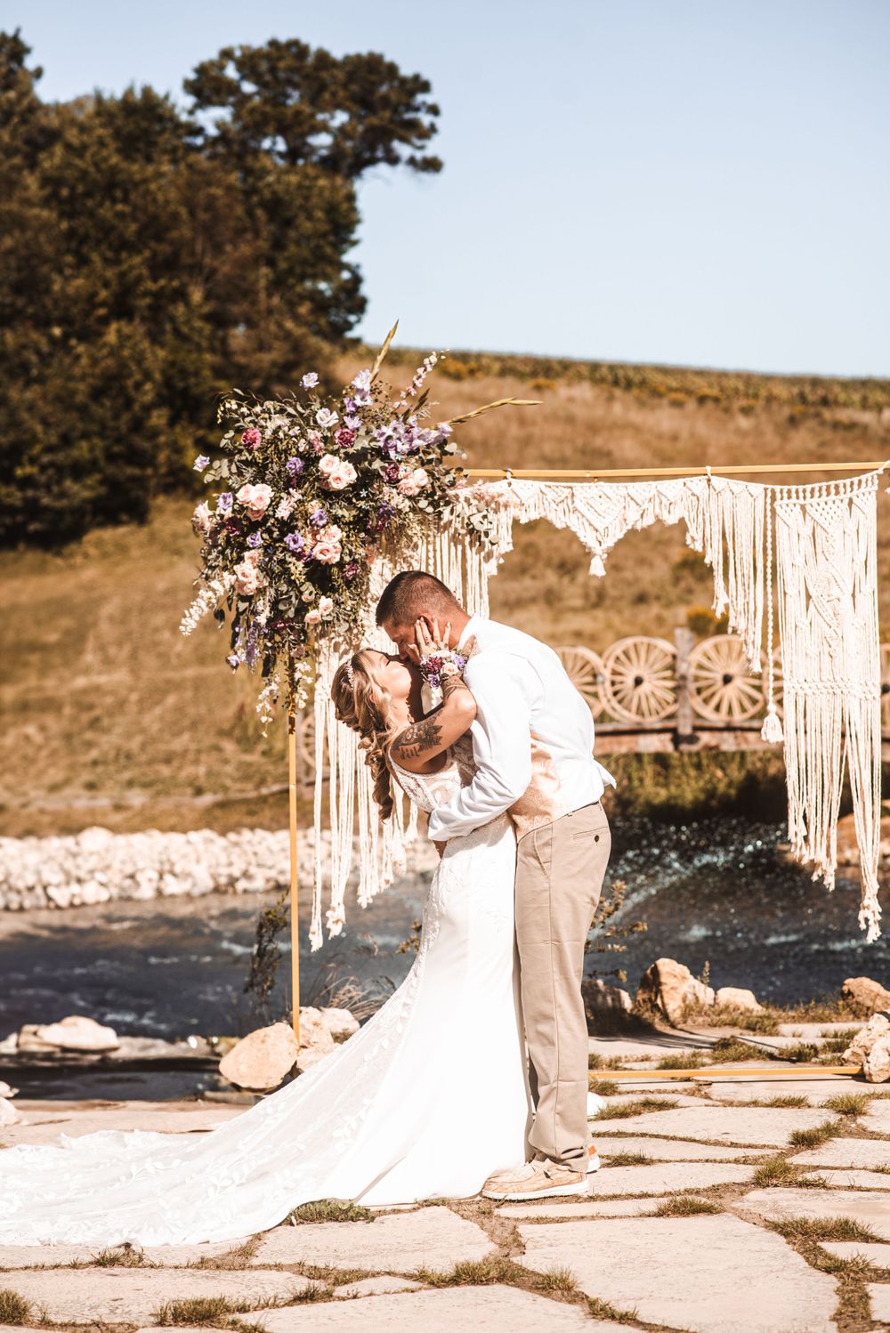 Couple kisses under floral arch with macrame backdrop at outdoor wedding.