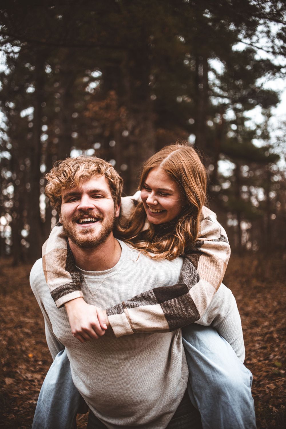 A couple shares a joyful piggyback moment in an autumn forest setting with pine trees in the background.