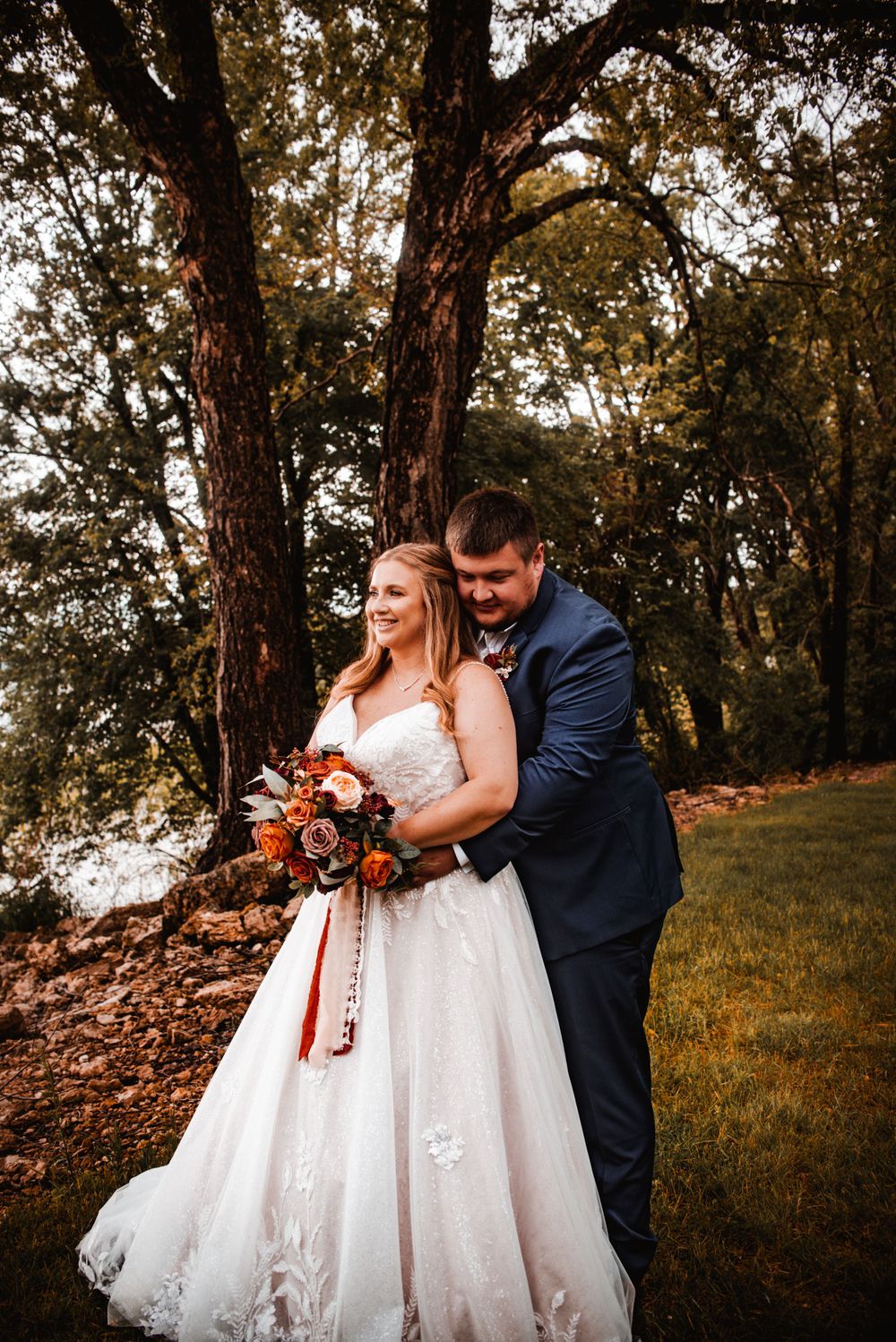 Autumn wedding photo of couple by trees with bride holding orange and burgundy bouquet.