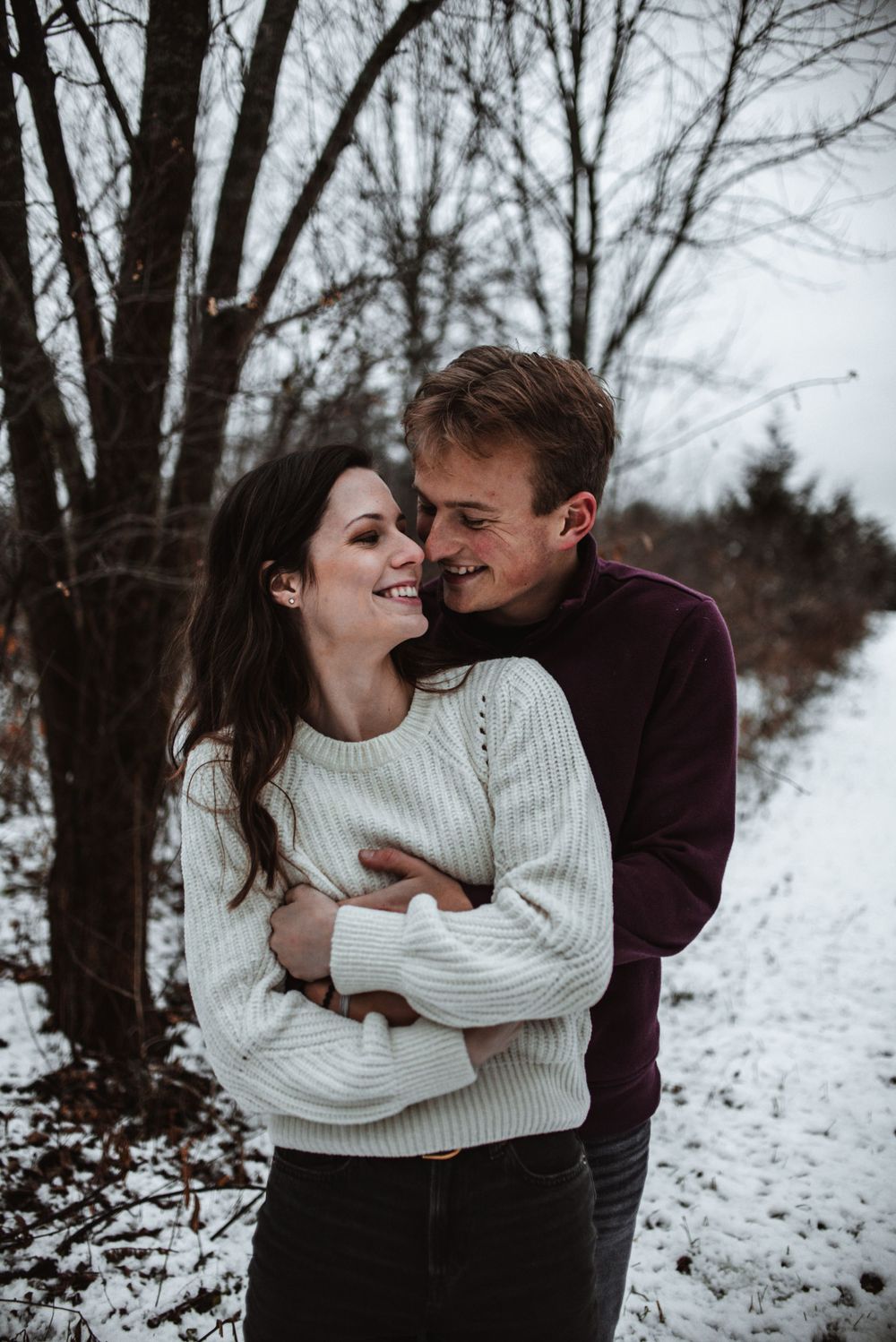 Romantic winter photoshoot of couple embracing on snowy path through bare trees at dusk.