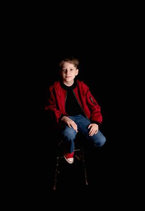 A child in a red jacket and jeans sits on a dark background in a series of progressive studio portrait poses.