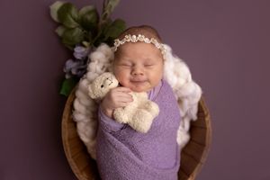 Sleeping newborn swaddled in purple wrap with teddy bear accessory in wooden bowl against mauve backdrop.
