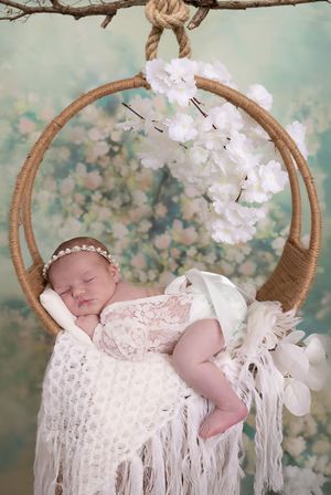 Sleeping newborn in white lace dress rests peacefully in rustic woven basket with floral backdrop.