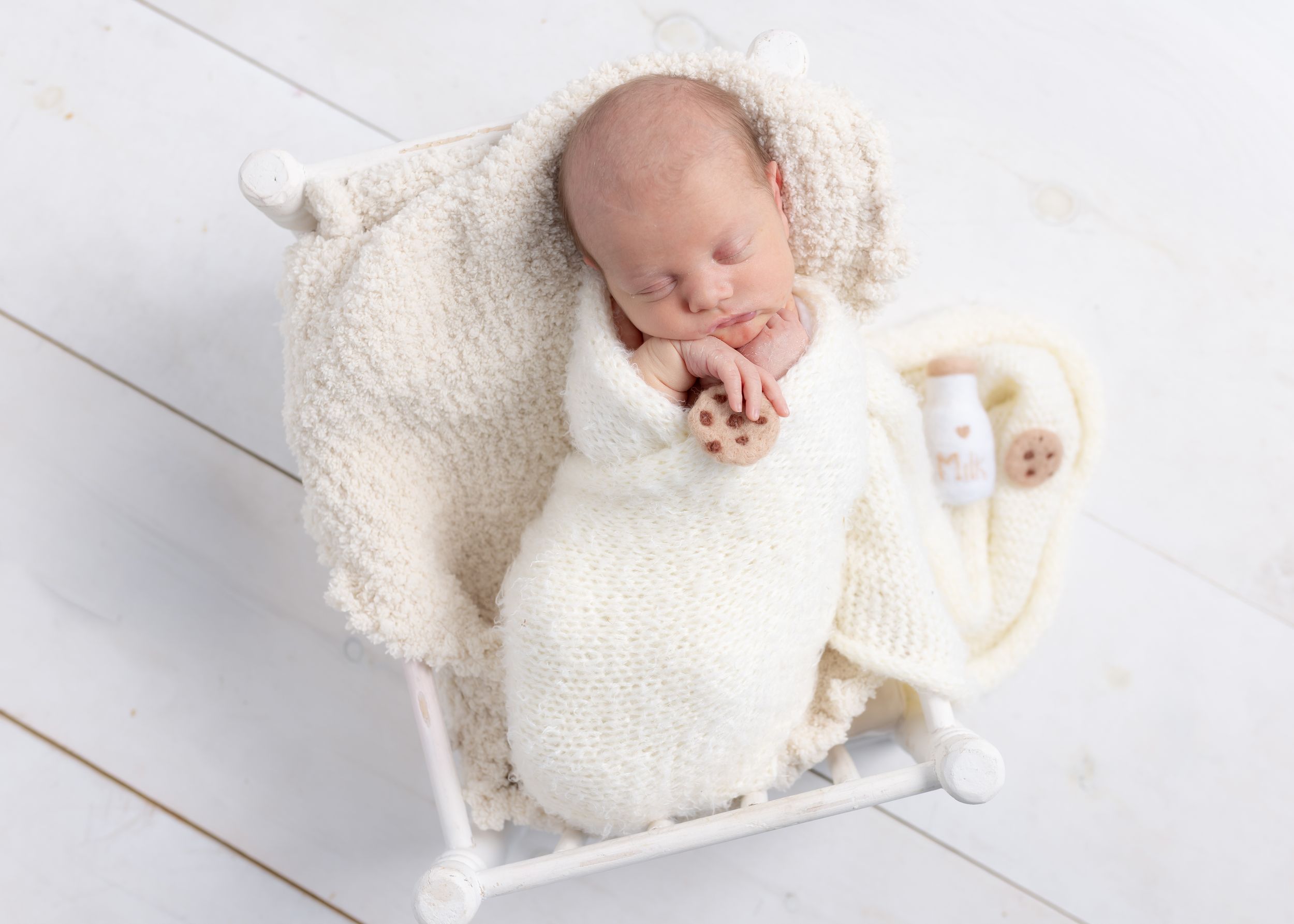 A sleeping newborn baby wrapped in soft white blankets lies peacefully on a white wooden floor.