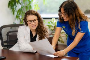 Two coworkers collaborate at a desk with papers and documents while discussing work details in an office setting.