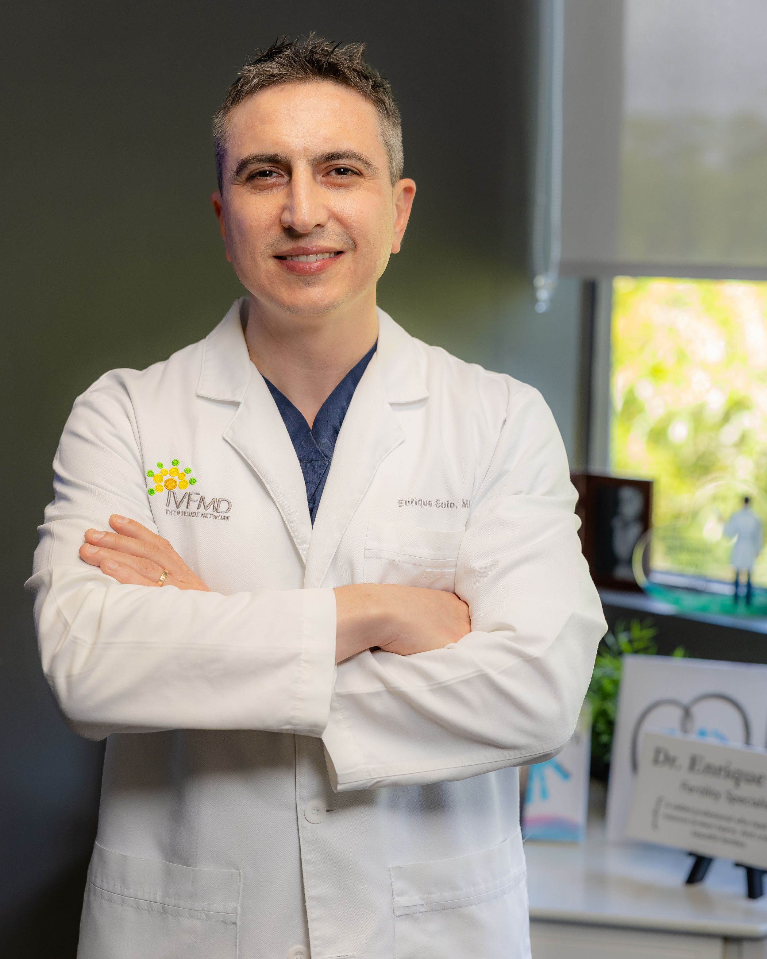 Medical professional in white coat with crossed arms standing in modern office setting with natural lighting.