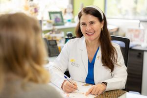 Healthcare provider in white coat and blue scrubs sits at desk writing notes during patient consultation.