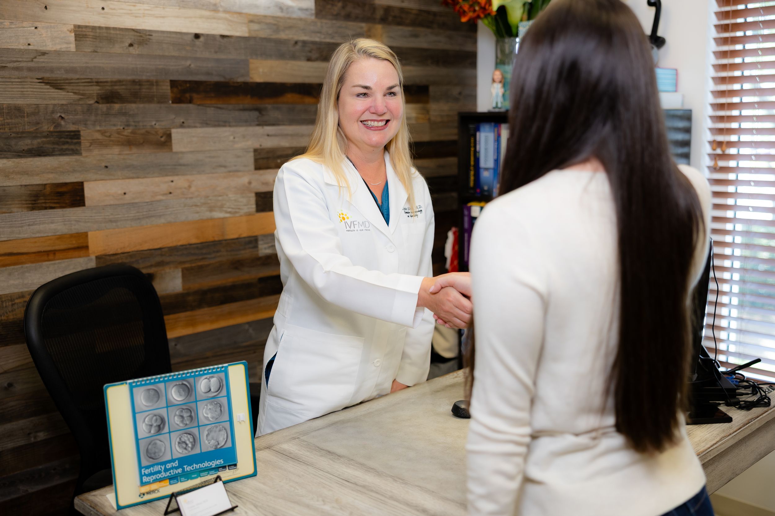 Two people dressed in white medical coats smiling and interacting in a modern office setting with wood-paneled walls.