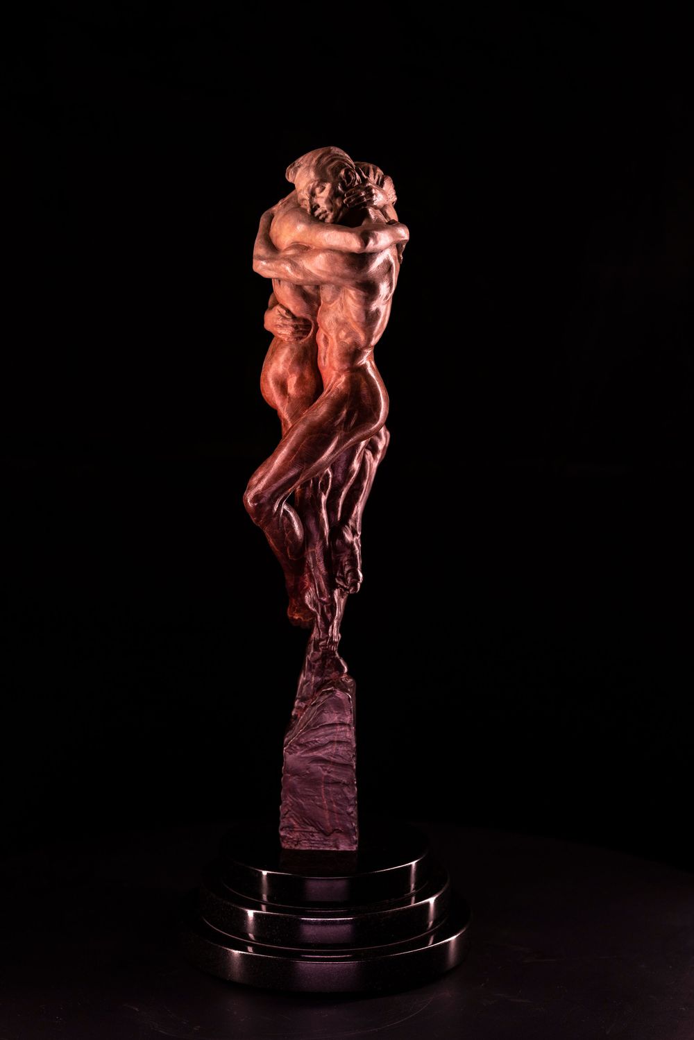 Bronze sculpture of entwined figures in dynamic poses displayed on dark pedestal against black background.