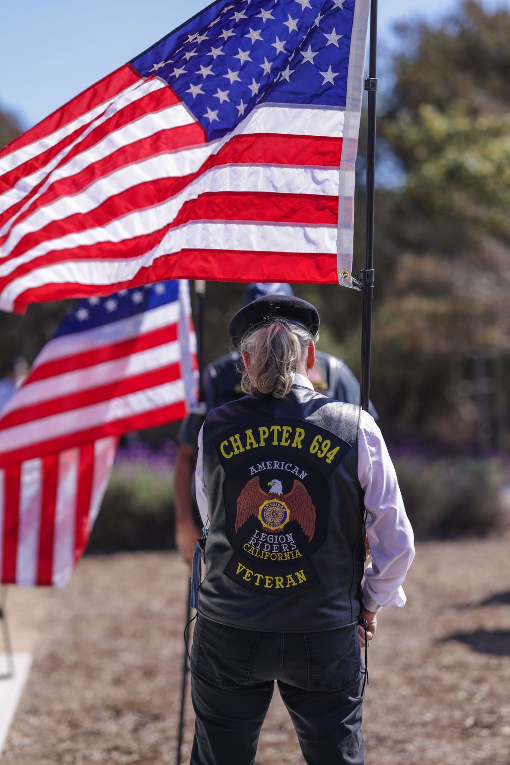 American flags wave in the wind as a veteran in a Chapter 891 vest stands in an outdoor setting.