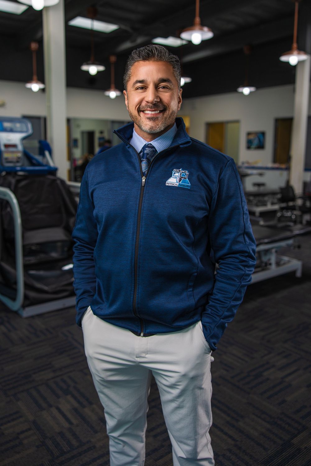 Professional wearing navy zip-up jacket and khakis standing in gym environment.
