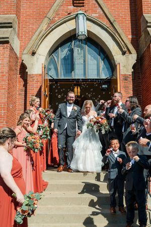 Wedding party exits historic brick church as guests celebrate with bubbles and cheers on the steps.
