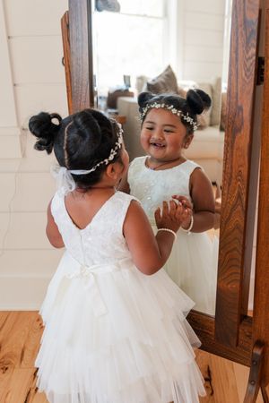Two flower girls in white dresses share a playful moment together in a sunlit indoor setting.
