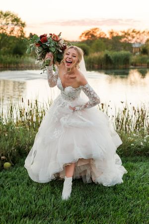 A joyful bride in a flowing white dress sits and laughs by a serene lake during golden hour.