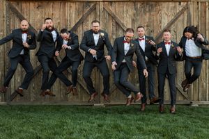 Groomsmen in dark suits perform synchronized jumping poses against a wooden fence and grassy backdrop.