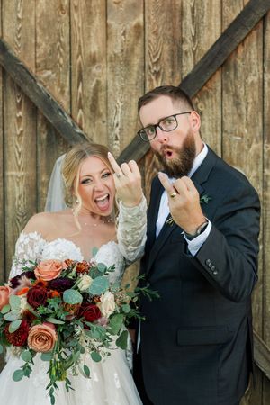A joyful wedding couple shares a candid laughing moment against a rustic wooden wall backdrop with peach floral bouquet.