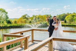A couple in wedding attire shares a romantic moment on a wooden dock overlooking a serene lake on a sunny day.