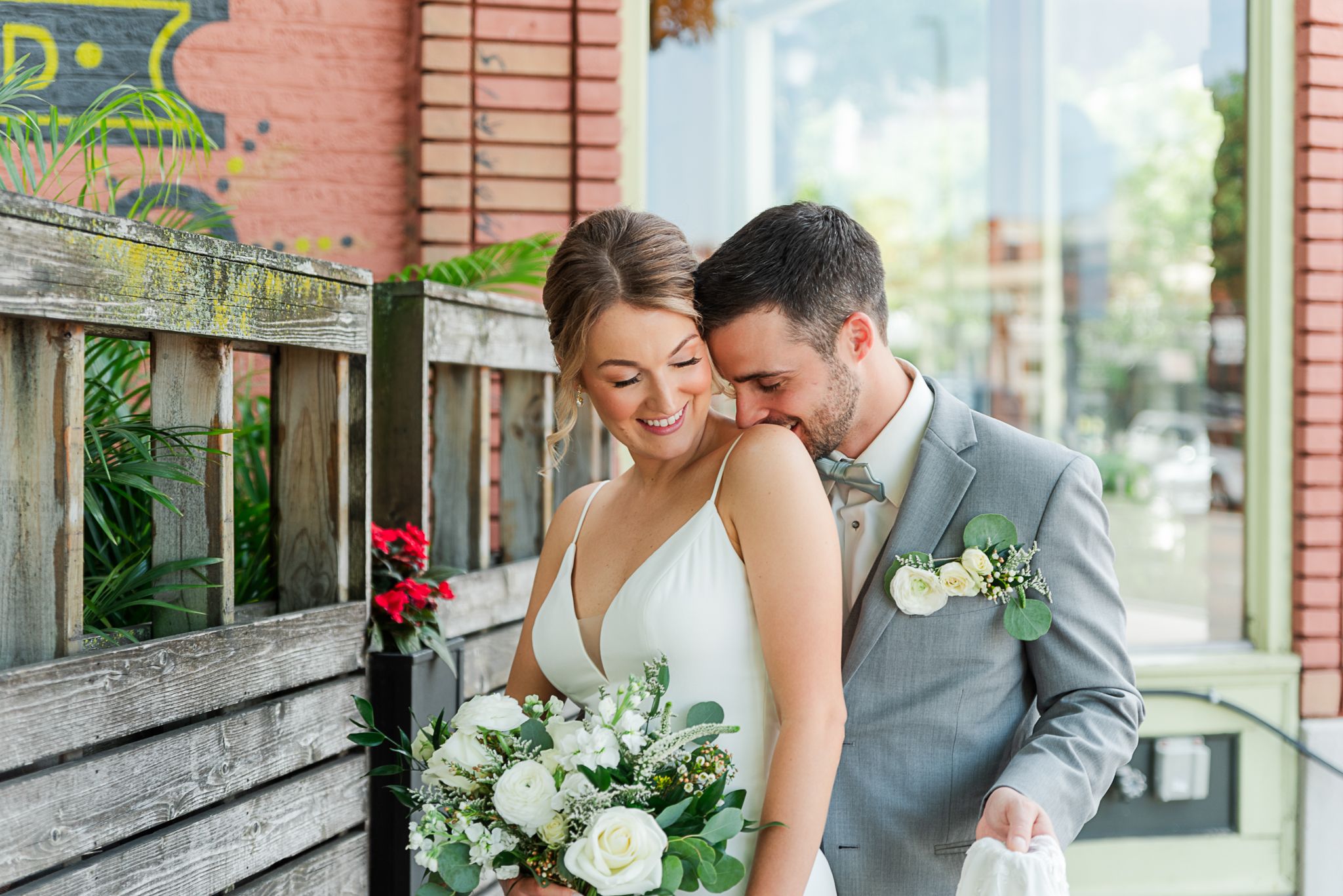 Sweet wedding couple shares an intimate moment on a wooden bench with white floral bouquet and red rose decor.