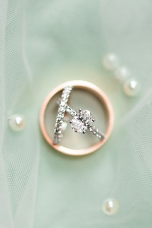 Elegant diamond engagement ring and wedding band displayed in a rose gold circular frame with scattered pearls on mint tulle.