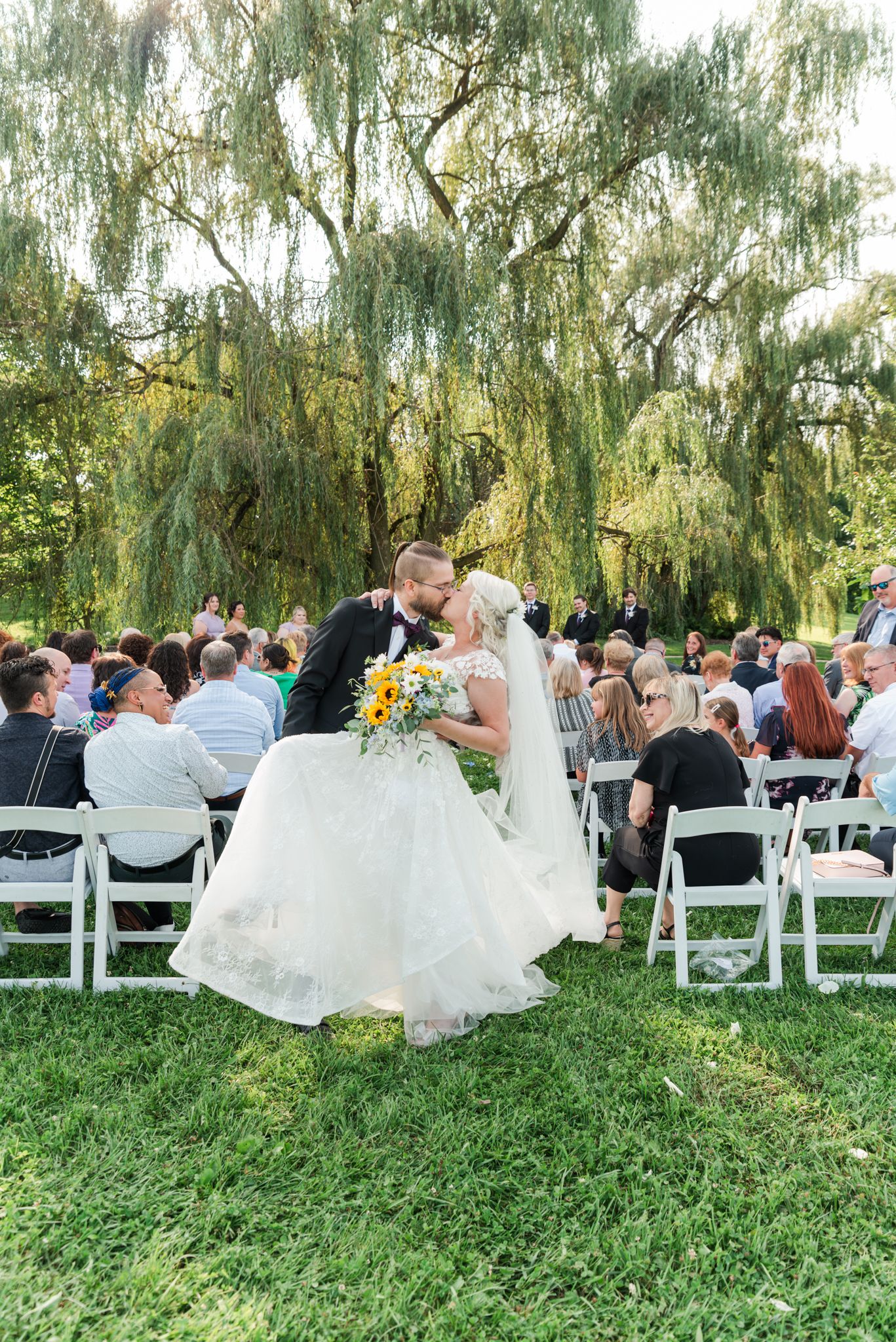 A wedding ceremony takes place under a majestic willow tree with guests seated on white chairs on the lawn.