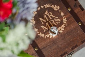 Rustic wooden ring box with engraved 'Mr & Mrs' text and floral details displayed against a blurred background.