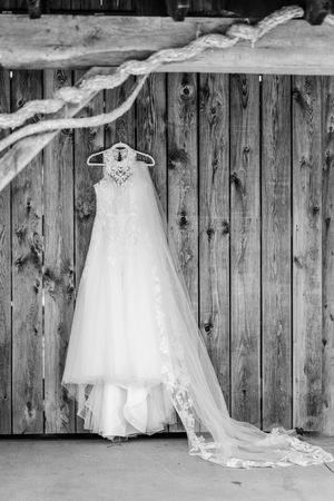 A white wedding dress hangs on a wooden wall in a rustic barn setting, displaying its elegant lace and tulle details.