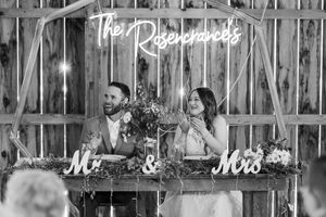 A black and white wedding photo showing a couple dancing together in a rustic barn venue with string lights overhead.