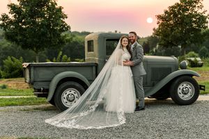 A vintage gray pickup truck serves as backdrop for a romantic sunset wedding photo with a flowing lace veil.