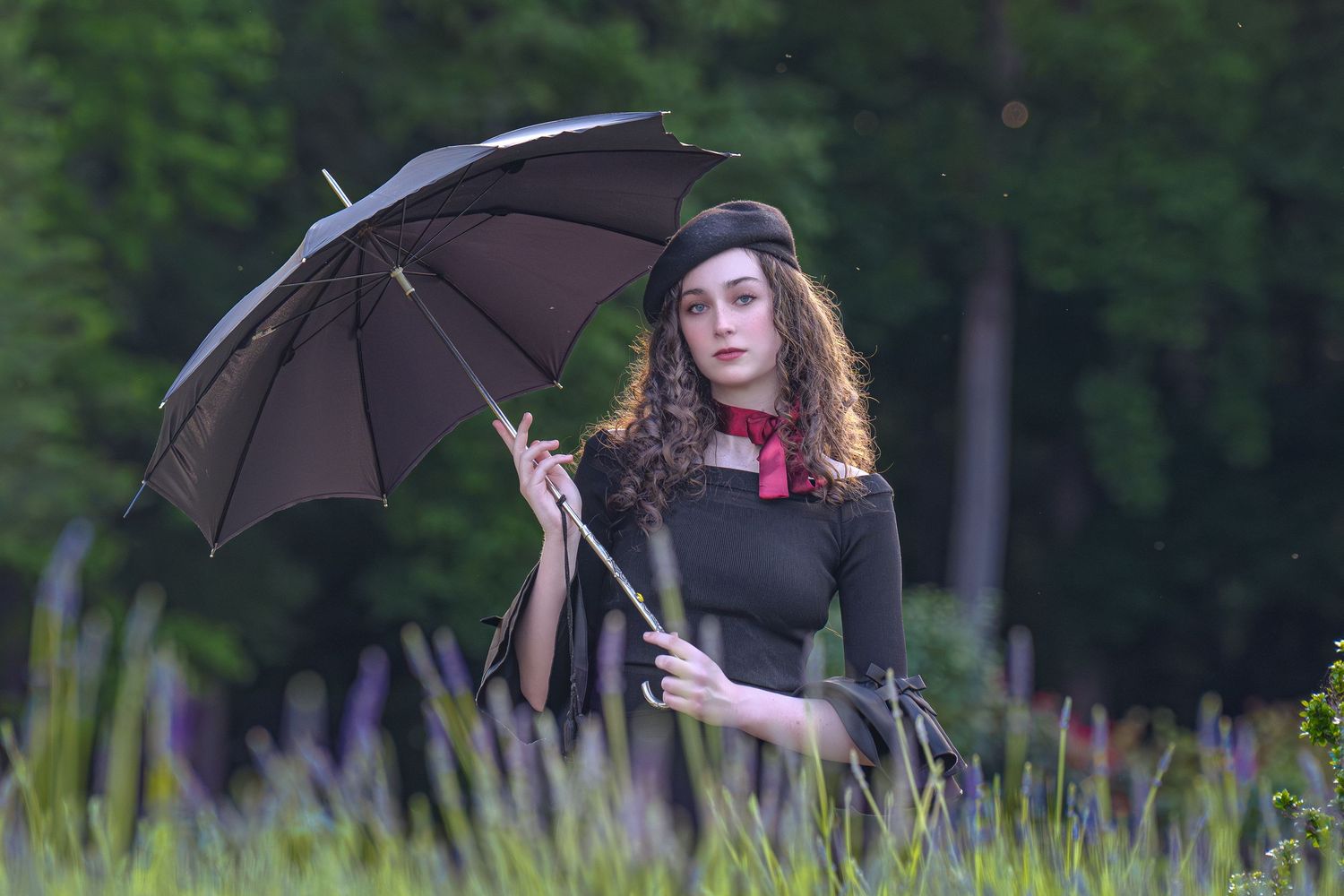 Young woman in black dress and beret holding a dark umbrella outdoors among tall green plants.Best photographer Williamsburg VA